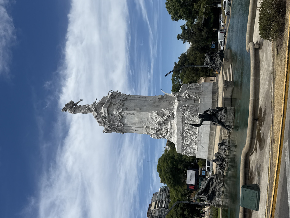 The image displays the Monument to the Two Congresses in Plaza del Congreso, located at the intersection of Avenida Rivadavia and Callao in Buenos Aires, Argentina, under a clear blue sky with scattered white clouds. The central subject is a tall, multi-tiered white marble monument featuring numerous relief sculptures and crowned by a standing allegorical female figure with outstretched arms. The base of the monument is encircled by a large, shallow fountain containing dark bronze sculptural groups, including a prominent male figure holding a trident, seated on a rock in the foreground water. Surrounding the monument are green trees and urban infrastructure. In the background to the left, multi-story buildings are visible, along with a billboard displaying "NETFLIX" and "YO LO CREO." Paved roadways with several white vehicles are present around the monument and fountain, indicating an active urban environment. The foreground shows a paved sidewalk area with low green foliage. No individual people are distinctly visible or engaged in specific actions.