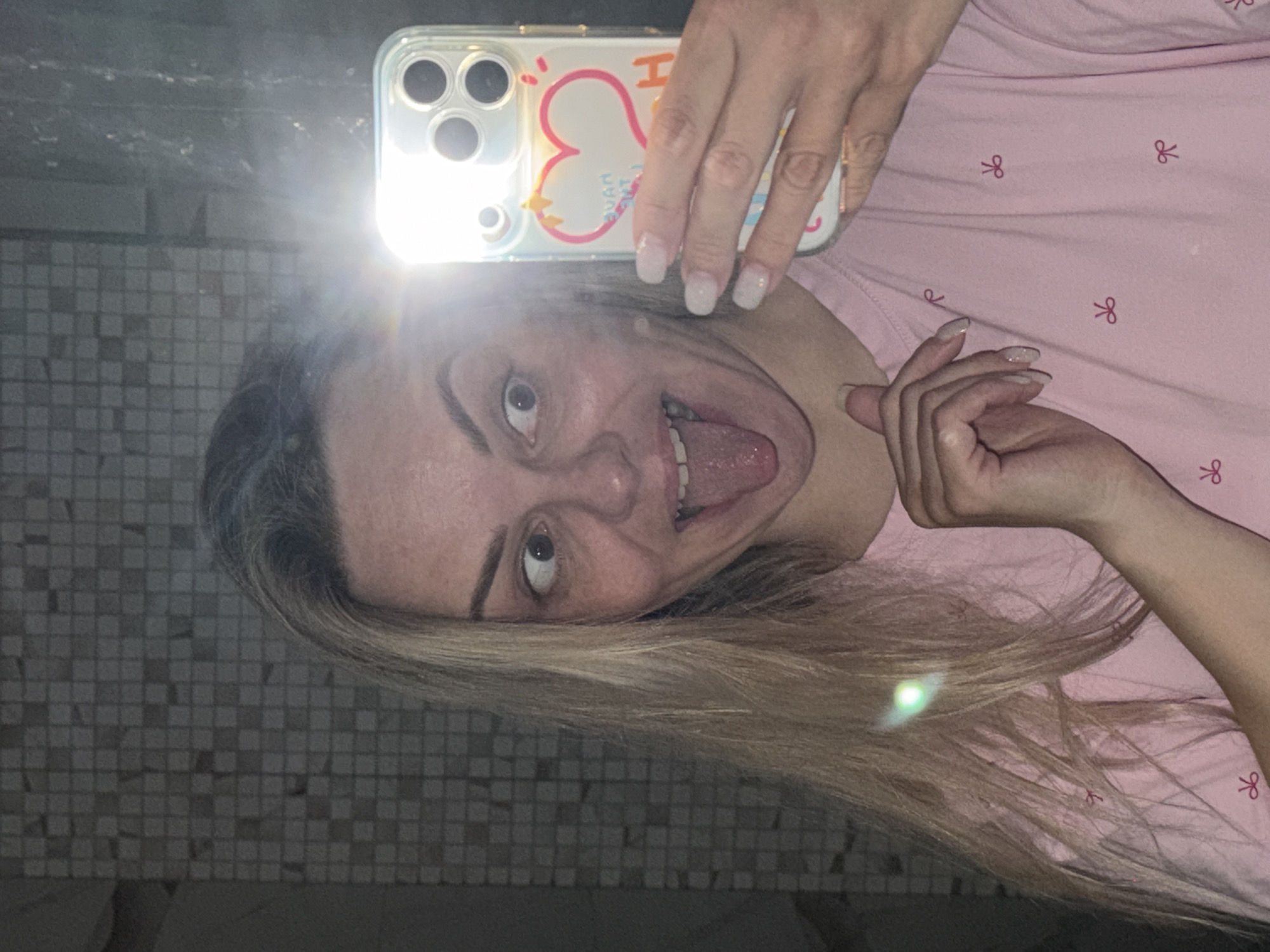 A woman is taking a selfie with her phone in what appears to be a bathroom, judging by the tiled wall behind her. She is wearing a light pink pajama top with a bow pattern. She is sticking her tongue out. The phone has a clear case with a white background and a cartoon heart design. The flash of the phone is reflecting brightly. The location in Votkinsk, Russia is unknown.