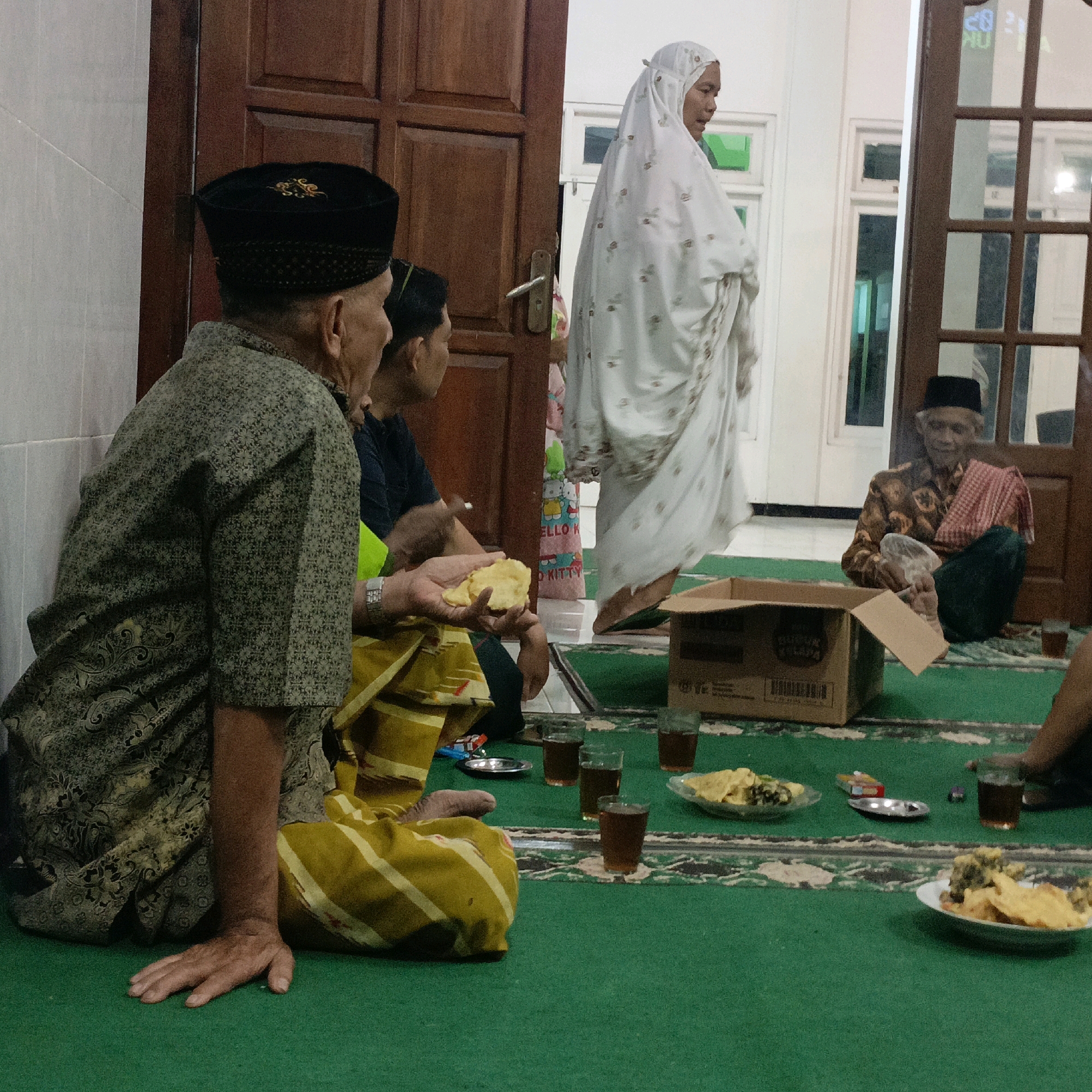 The image captures an indoor scene, likely a home in Salatiga, Indonesia, where a group of people are gathered on a green carpeted floor. In the foreground, a man wearing a black peci hat and a patterned batik shirt sits with his back mostly to the camera, his left arm resting on his lap. To his right, another man in a dark shirt is partially visible. A woman, dressed in a white hijab and a patterned white garment, stands in the background, her right arm slightly raised. Further to the right, an elderly man in a patterned shirt and a red and white checkered sarong sits with his legs crossed, holding a white cloth.
The floor is covered with a green carpet featuring an ornate pattern. Scattered on the carpet are several items: a brown cardboard box, small clear glasses with dark liquid (likely tea), plates of food (appearing to be snacks or fried items), and a small silver dish. The background shows wooden doors and white walls, with glimpses of a window with framed panes. The lighting suggests an evening setting, with soft illumination contributing to a relaxed, intimate atmosphere. The overall impression is of a casual gathering, possibly a family or community event, involving food and conversation.