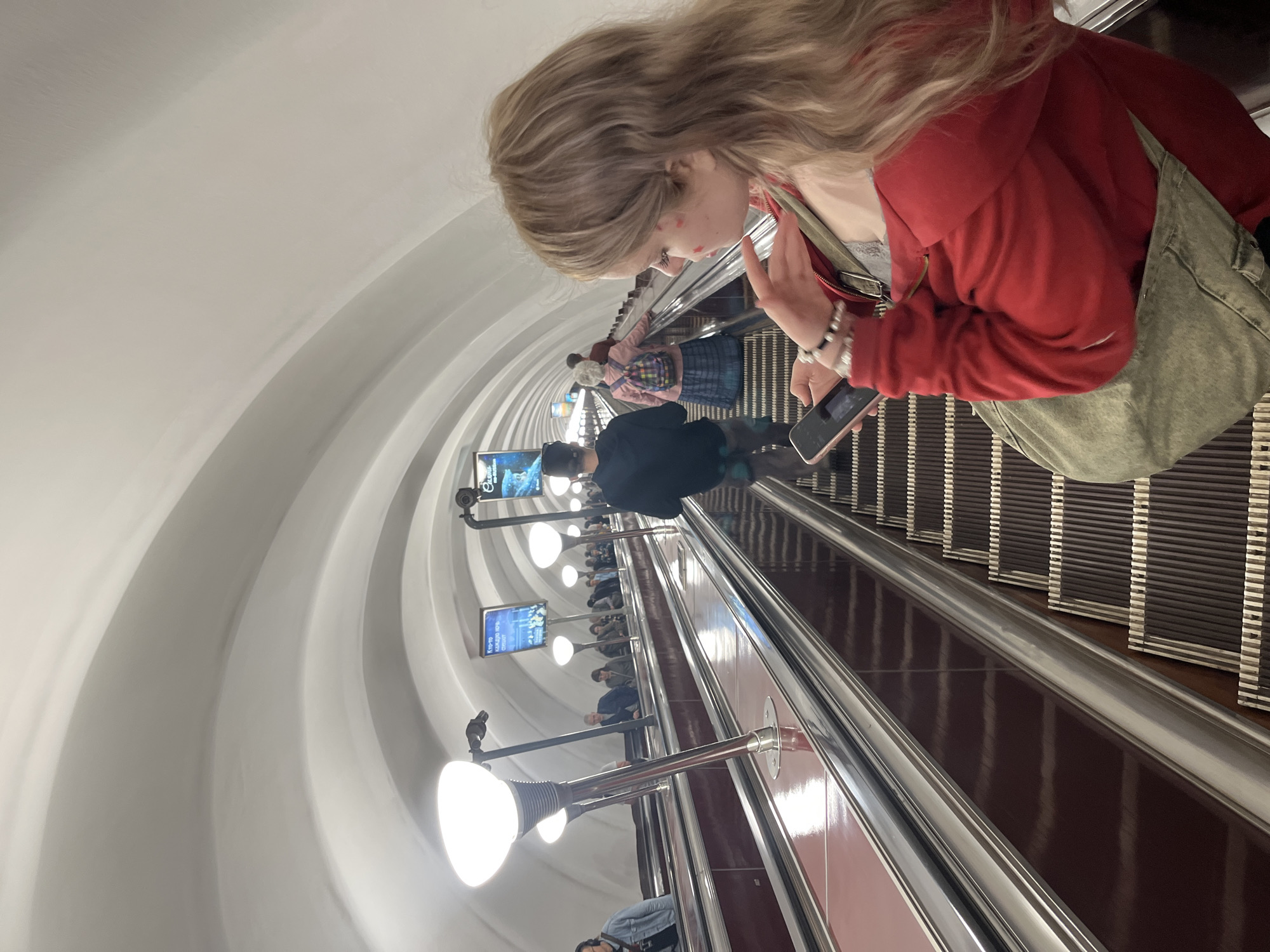 This vertically-oriented image captures a scene within a deep underground metro station, likely in Saint Petersburg, Russia, as people descend a long escalator. The tunnel features curved, light-colored walls and is illuminated by numerous bright, dome-shaped lights mounted on poles alongside the escalator.
In the immediate foreground, a young woman with long, wavy blonde hair is the central subject. She is looking down at a mobile phone in her hands and wears a red hooded jacket over a light-colored top, along with an olive-green cross-body bag. Notable details include red markings on her face (possibly makeup or small stickers) and a beaded bracelet on her wrist.
Behind her, several other individuals are also riding the escalator downwards. One person directly behind her, wearing a dark top and a baseball cap, is also looking at a phone. Further down, another person is visible in a pink jacket and a plaid skirt, carrying a colorful backpack.
On the left side of the escalator tunnel, two digital screens display advertisements or information. The upper screen shows what appears to be a movie poster with Russian text partially visible, including "Сказо..." (Skazo...) and "на экранах" (on screens). The lower screen also displays Russian text, including "Куда" (Where to), "Каждую среду" (Every Wednesday), and "Особая" (Special). The long escalator, deep tunnel, and visible Russian text strongly suggest the Saint Petersburg Metro. The time of day is indeterminate due to the underground setting.