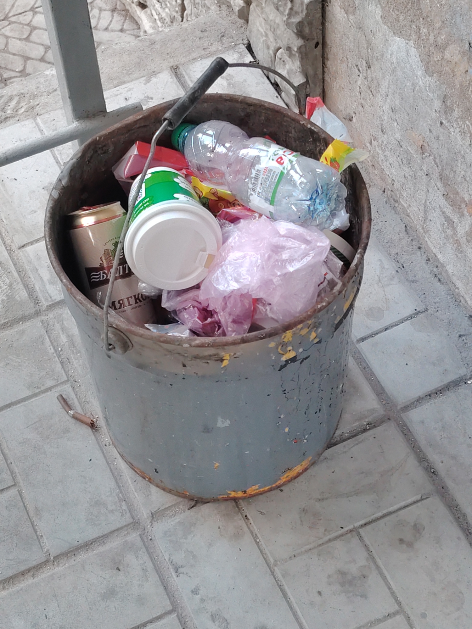 A metal bucket filled with various refuse, including plastic bottles, cans, and plastic bags, is situated outdoors on a tiled pavement. The bucket is gray with orange and yellow paint chips, and it has a black handle. To the left of the bucket, a metal pole and stone structure are visible. The ground beneath the bucket is composed of light gray square tiles. The scene appears to be in an urban setting, possibly in Nizhny Novgorod, Russia, given the mention of the city.