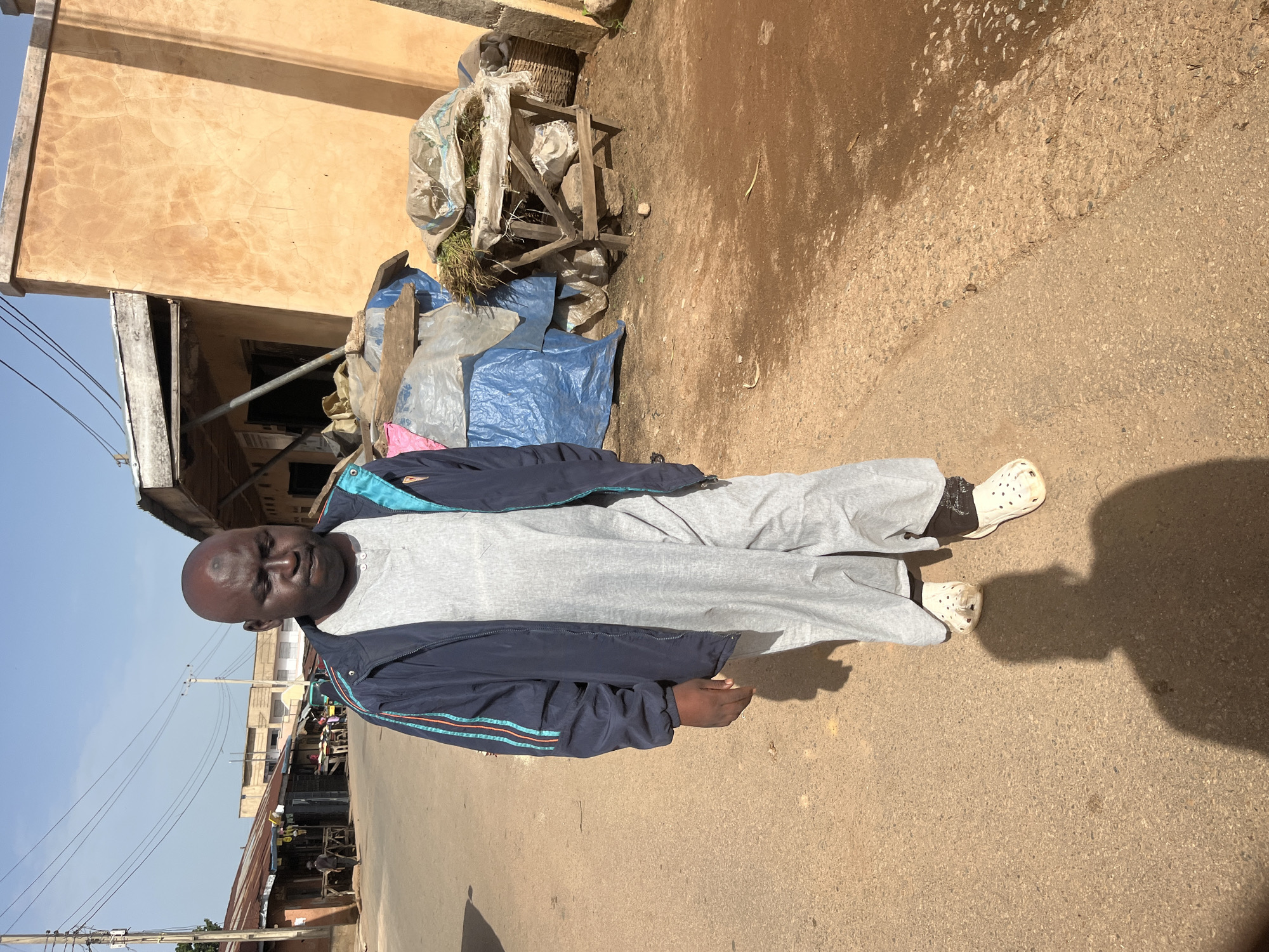 A man stands on a dirt road in what appears to be a market area. He is wearing a light grey t-shirt, grey sweatpants, a dark blue jacket with teal and red stripes on the sleeves, and white Croc-style shoes with black socks. He has dark skin and is bald. The man's expression is neutral.
Behind him, a building with a textured, light-colored wall is visible. To the left of the building, there are some structures with corrugated metal roofs, suggesting a marketplace. Various objects are piled up in front of the building, including blue tarps, plastic bags, and what looks like bundled vegetation. A wooden structure, possibly a cart or shelving unit, is also present among the piled items.
The ground is a mix of dirt and asphalt, with some areas appearing wet or muddy. Sunlight casts a shadow of the man on the ground, indicating it is daytime. The sky is clear and blue. In the background, more market stalls and buildings are visible, along with utility poles and wires, suggesting an urban or semi-urban setting. The overall scene conveys a sense of an outdoor market environment in Nigeria, likely Jos, given the location context. No visible text is present in the image.