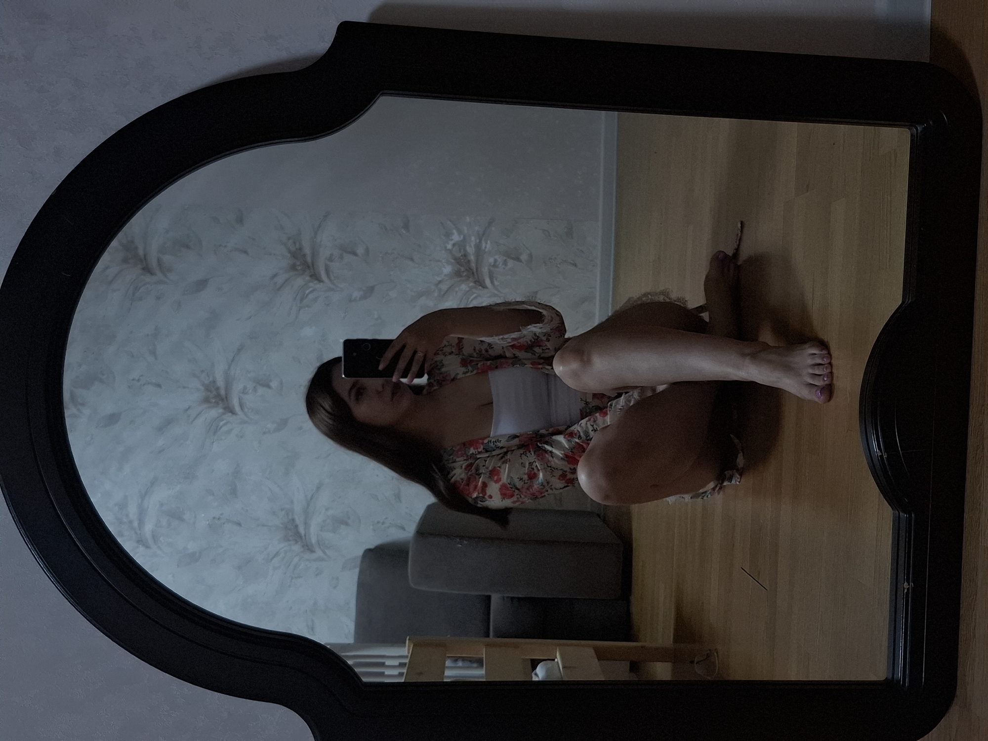 A mirror reflects a woman sitting on a wooden floor, taking a selfie. The woman wears a floral robe over a white top. Her reflection includes wallpaper with a patterned design and a grey couch. The mirror's frame is black and ornate. A location detail provided is Chaikovsky, Russia.