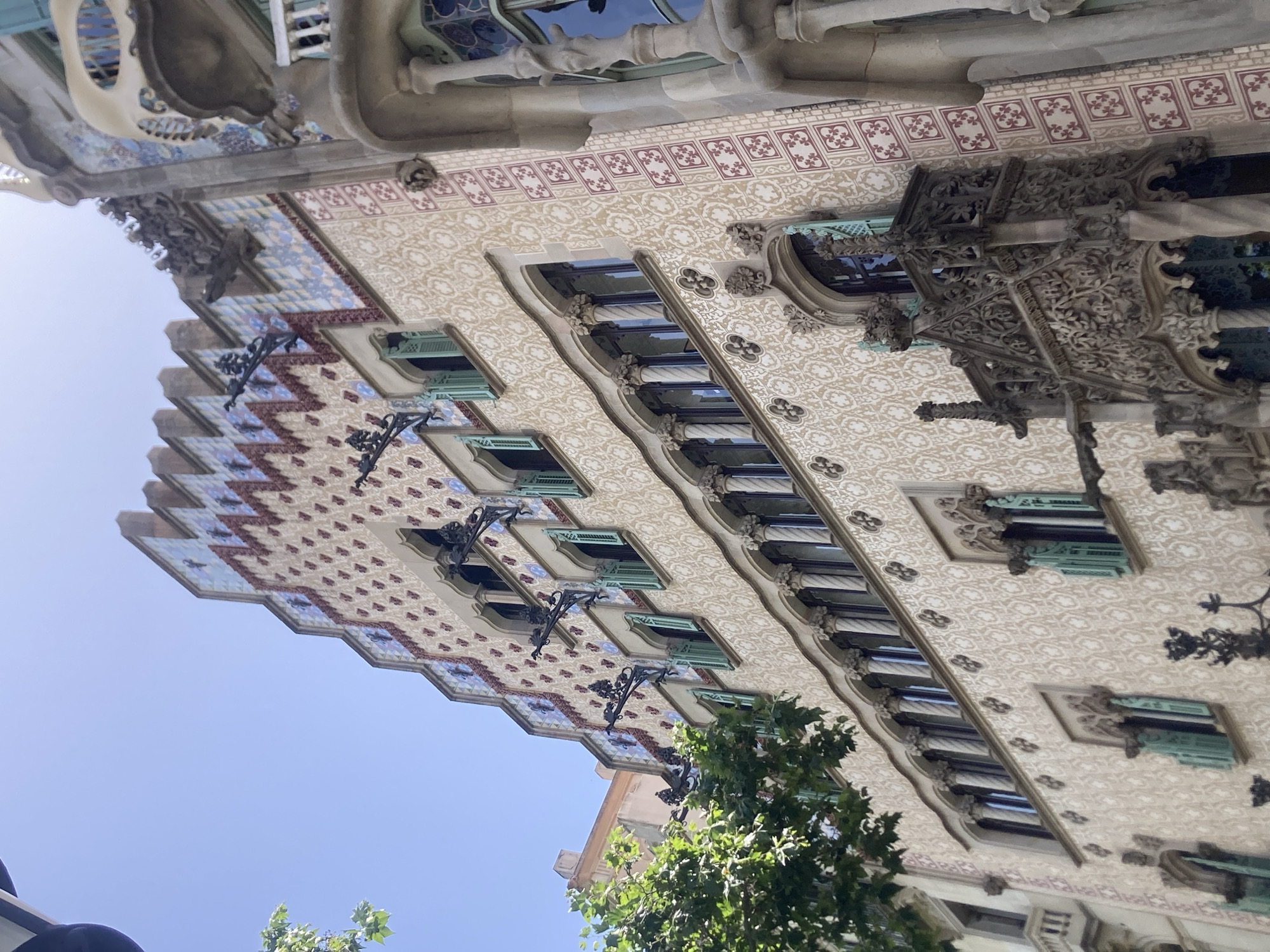 This is an image of a building with ornate architectural details, possibly taken in Barcelona, Spain. The building's facade features a decorative tiled pattern in shades of beige, red, and blue, with repeating floral motifs. There are numerous windows with green shutters, recessed into the facade and framed by intricate stonework. Decorative elements in sculpted stone are also visible, including what appear to be gargoyles or grotesques. The perspective is from below, looking upwards, with a clear blue sky in the background, suggesting it is daytime and the weather is fair. A tree with green leaves is partially visible in the foreground, adding a natural element to the scene. The overall impression is one of a historic and elaborately designed building.