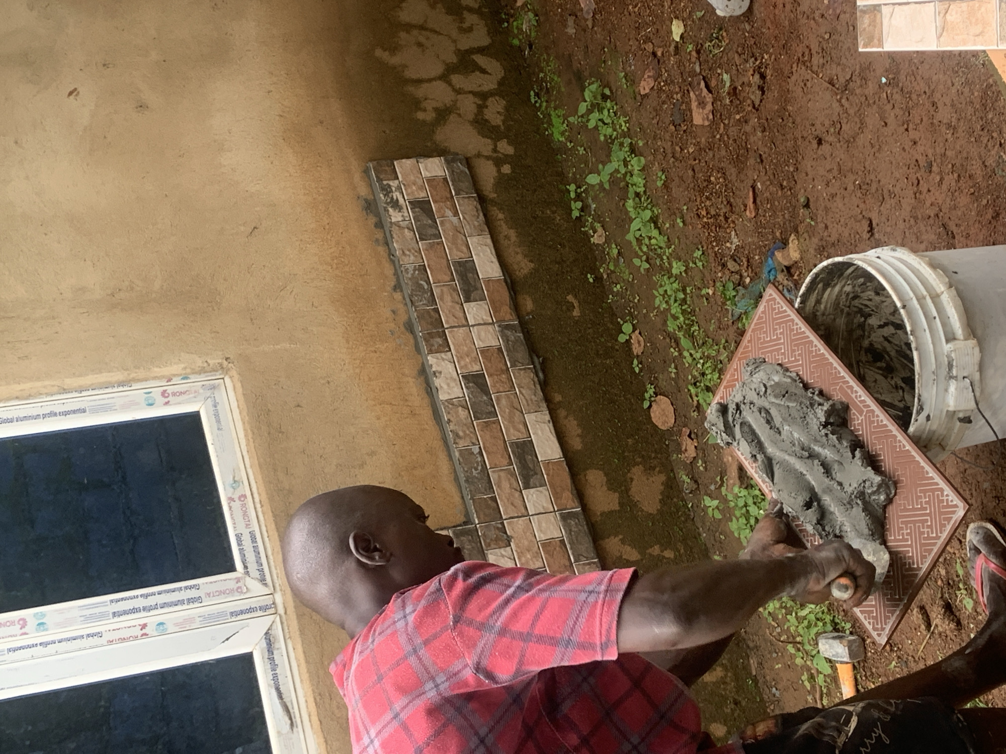 In Agban, Nigeria, a man is shown tiling an exterior wall. He is wearing a red plaid shirt and dark shorts, with his head bowed as he works. In his hands, he holds a trowel, applying gray mortar to a ceramic tile. The tile itself has a decorative pattern. The wall being worked on is a light brown stucco, with a section already tiled in a brick pattern of browns and creams. Below the wall, the ground is dirt with some green vegetation. To the left, a white bucket sits on the ground, and a hammer is visible. A window with white trim is in the upper portion of the image.