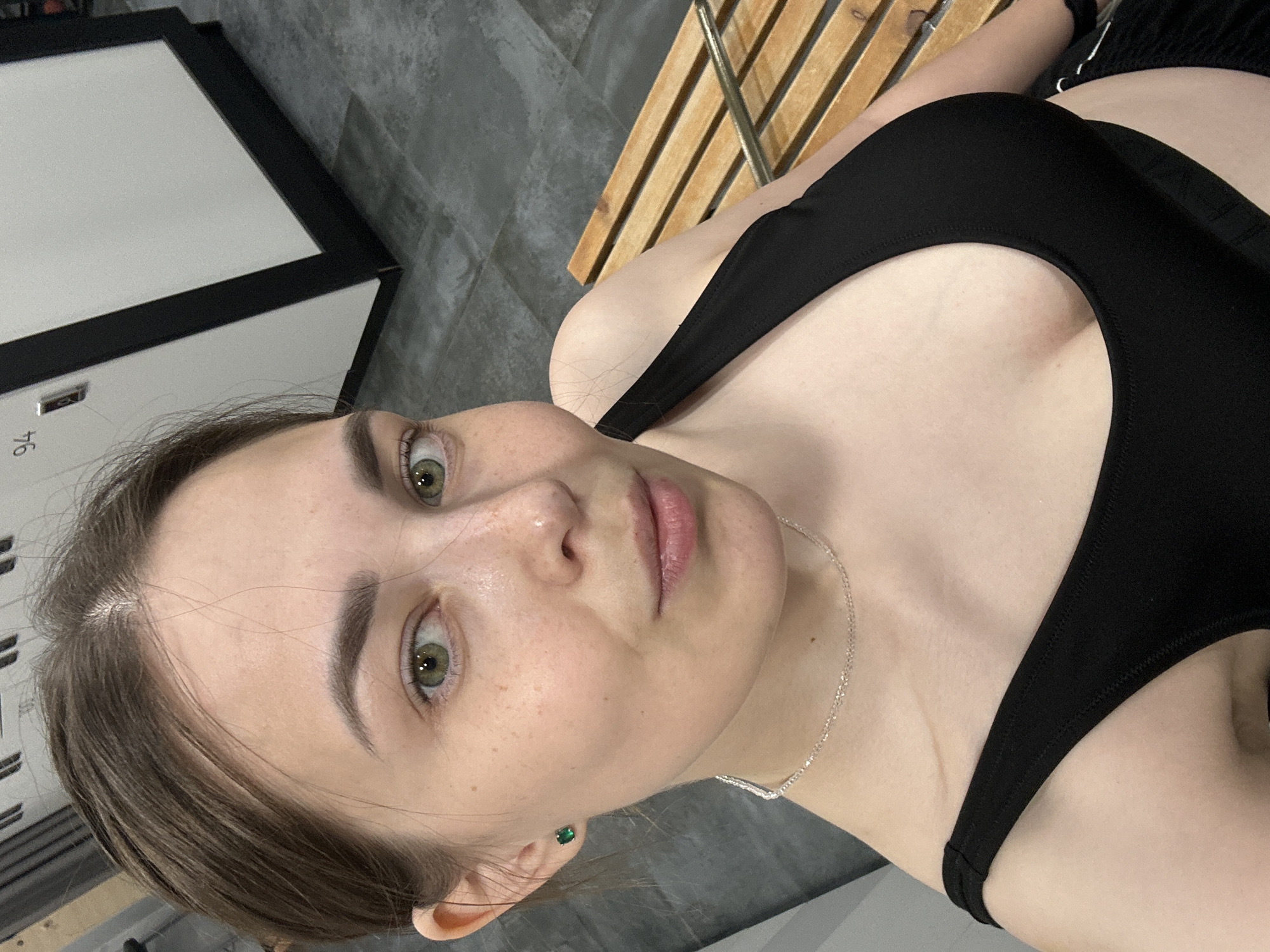 A young woman with fair skin, brown hair pulled back, and green eyes is taking a selfie in what appears to be a gym locker room. She is wearing a black sports bra and a silver necklace. She has a small green earring in her left ear. Behind her, there are lockers with the number 94 visible on one of them, and a wooden bench. The floor is tiled. The scene suggests a casual, personal moment in a public facility. The overall impression is one of youthful self-awareness and a focus on personal appearance.