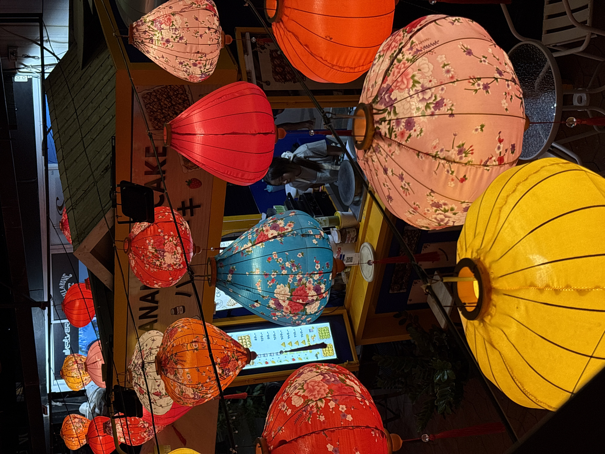 The image depicts an outdoor scene at night in Choeng Thale, Thailand, showcasing a vibrant display of illuminated paper lanterns. The lanterns are of various shapes, sizes, and colors, including red, orange, yellow, blue, and pink, many adorned with floral patterns. They are suspended by wires, creating a festive atmosphere above a food stall.
The food stall appears to be a crepe stand, with a young woman in a grey apron visible behind the counter, preparing food. A menu board with Thai script and prices is displayed prominently on the stall. In the background, a building with a sloped, dark-colored roof and signage that includes the words "SILVER" and "BILLABONG" is visible.
The foreground is dominated by several large, brightly lit lanterns. A large yellow lantern occupies the bottom left of the frame, and a pink floral lantern is positioned to its right, partially obscuring a glass-topped table and chairs. Other lanterns, including red, orange, and blue ones with floral designs, are scattered throughout the midground.
The ground is paved, and some greenery is visible in the background. The overall impression is one of a lively night market or street food scene.