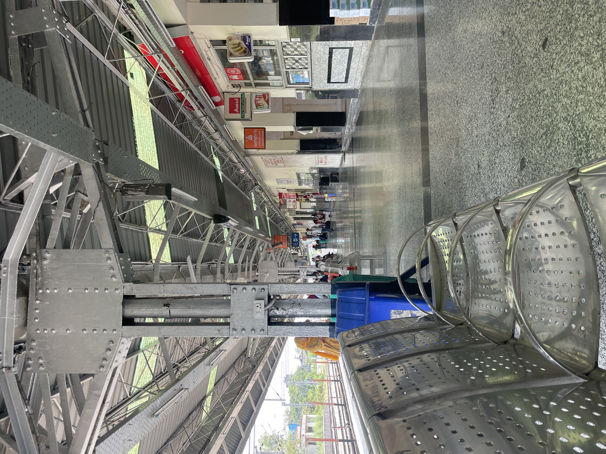 The image depicts an interior view of a covered train station platform. In the foreground, several metallic benches with perforated seating are visible. To the left, a blue trash bin and a green trash bin are positioned next to a support pillar. The pillar itself is metallic and shows some signs of wear.
The platform floor is made of speckled grey terrazzo. Above, a complex network of steel beams and corrugated metal roofing creates a canopy over the platform. Fluorescent lights are installed along the underside of the roof structure.
In the middle ground, the platform stretches into the distance, lined with shops and stalls. Signs are visible, including one for "Amul The Taste of India" and another with Hindi text that appears to be advertising an "Executive Lounge." A woman in a bright yellow and orange garment is standing near the edge of the platform on the left side of the image, looking towards the tracks. Other individuals are scattered throughout the platform, some walking and others seated.
The background shows train tracks and some greenery beyond the station structure. The overall environment is that of a functional, public transportation hub. This scene is consistent with the appearance of the Pathankot Railway Station in Pathankot, India, which is known for its covered platforms, shops, and seating areas.