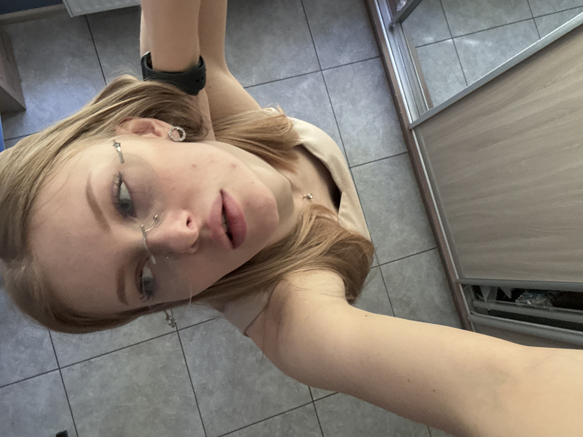 A young woman with light blonde hair and fair skin is positioned in the center of the frame, angled towards the camera in a selfie-style shot. She is wearing glasses and has multiple piercings on her face, including nose and ear piercings. A black smartwatch is visible on her left wrist. She wears a light-colored top.
The background consists of gray floor tiles and a light brown sliding wardrobe door.
The location is indoors, within a building in a city (Perm, Russia is specified, but this is not directly evident from the image alone).