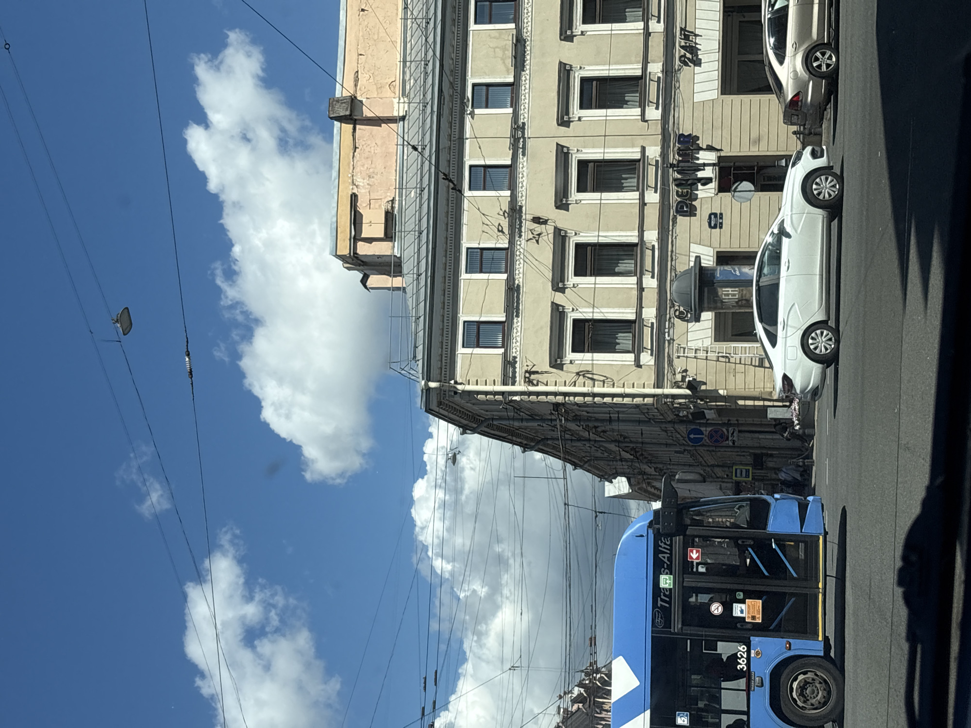 This media file captures an urban scene in Saint Petersburg, Russia, on a bright, cloudy day, likely midday given the shadows. The primary subjects are a blue bus with the text "Trans-Alfa" visible on its side and a white car parked on the street. The bus has the number "3626" printed on its front wheel arch.
The scene unfolds along a street lined with historic European-style buildings. The architecture features multiple stories with numerous windows, some adorned with decorative frames. The buildings show signs of age, with some plasterwork appearing worn or faded. Overhead, a complex network of electrical wires crisscrosses the sky, indicating a common urban infrastructure element. In the background, more buildings and street elements are visible, including traffic signs and a street lamp.
The weather appears to be a mix of sun and clouds, with large white cumulus clouds prominent against a bright blue sky. The lighting suggests a sunny period, casting distinct shadows on the road and buildings. There is no discernible activity or event taking place beyond typical street life, with vehicles and buildings suggesting a functional urban environment. The overall impression is one of a typical day in a historic European city, with a focus on the street-level activity and the architectural backdrop.