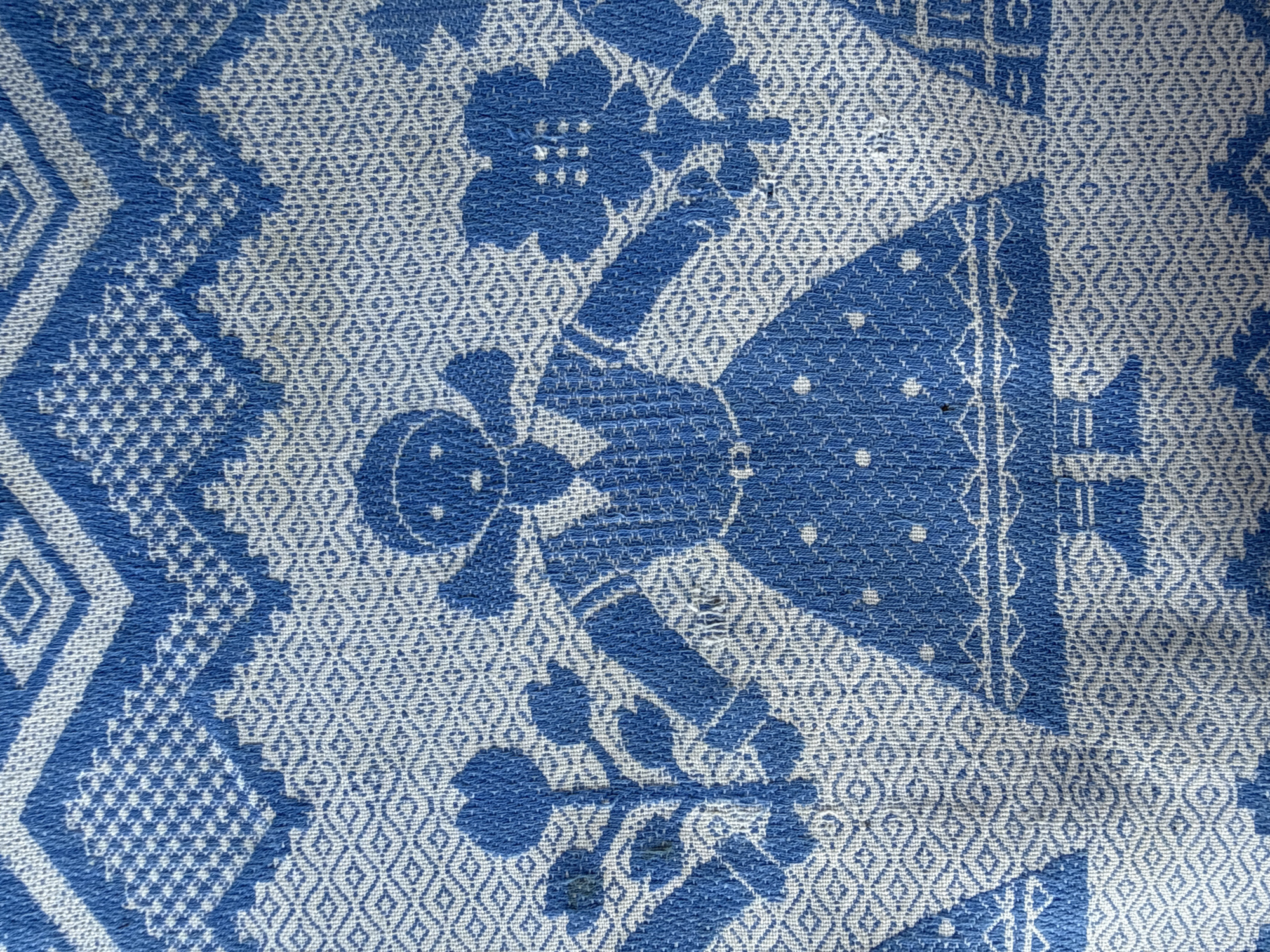 This is a close-up, eye-level shot of a blue and white patterned fabric, possibly a tapestry or woven cloth. The pattern features stylized figures, geometric shapes, and floral motifs. In the center, a human-like figure in a dress stands with arms outstretched, holding a flower in each hand. The figure has a round head with simple facial features and a bow tie. To the left and right of the central figure are depictions of plants with multiple leaves and flower heads. Above the figures, there are zigzag patterns and diamond shapes. Below the central figure, there are indications of other elements, possibly houses or more stylized figures. The fabric appears to be textured and slightly rumpled, with some visible wear or damage, including small holes. The image does not provide any information about the city of Малдыкасы, Russia, or any specific location within it.