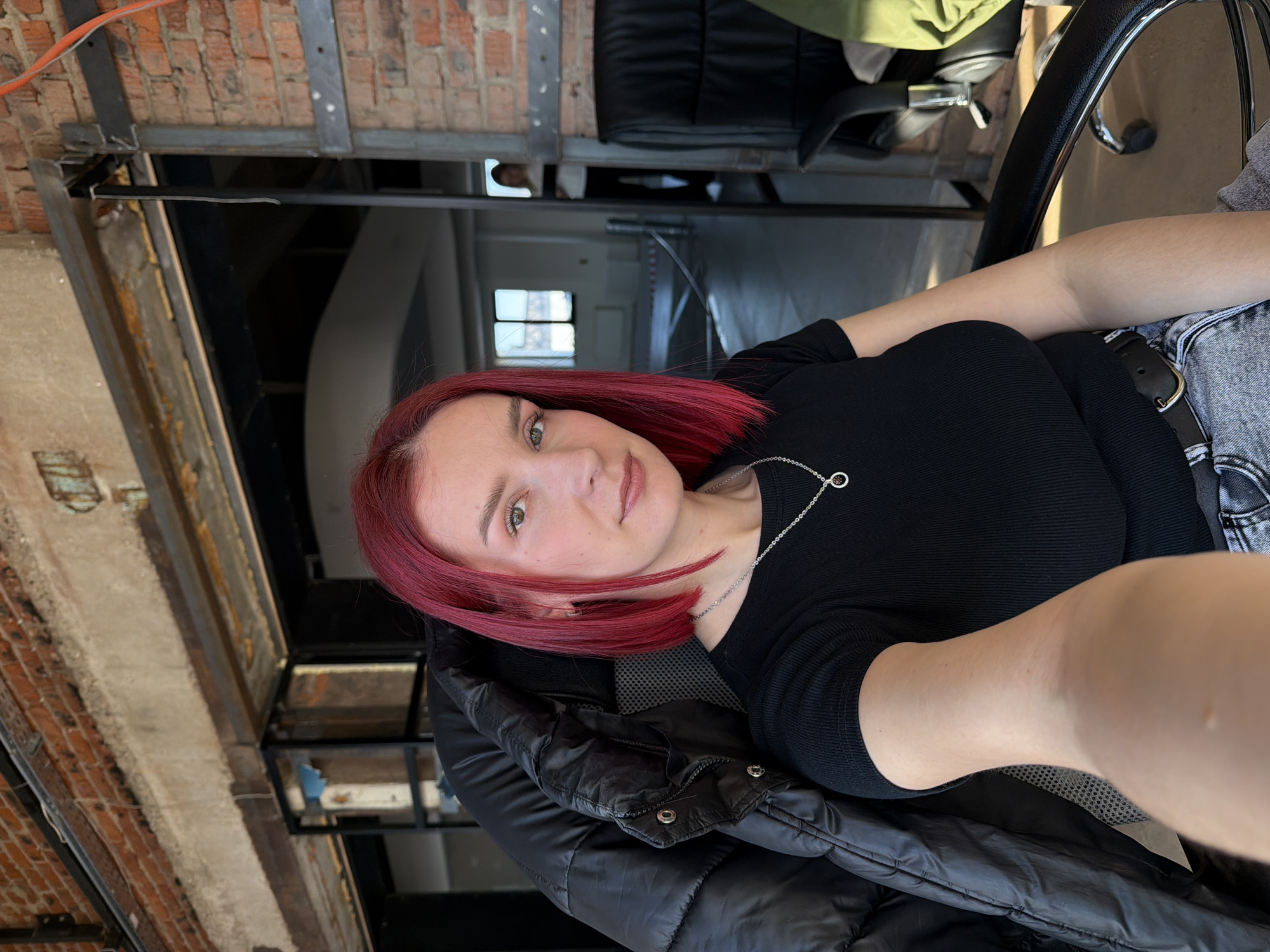 A woman with red hair and green eyes sits in a black chair, wearing a black top and jeans, a black jacket is draped over the chair. She is taking a selfie. The background shows a room with brick walls and metal beams, and a window in the distance. This may be a building in Chaikovsky, Russia.