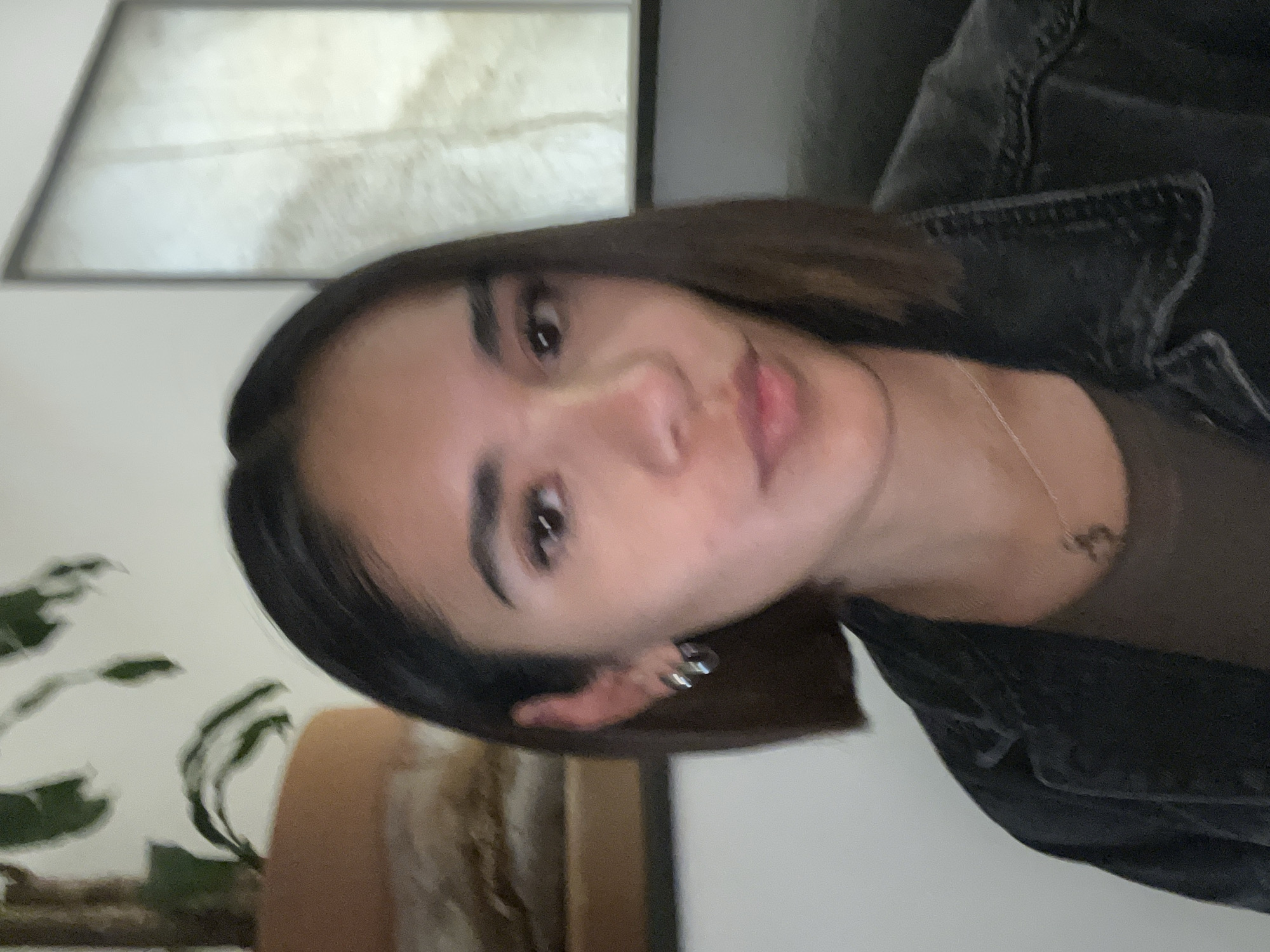 A person is facing the camera indoors. The person is wearing a dark jacket, a brown shirt, and earrings. There's artwork on the wall behind the person, and part of a houseplant is visible on the left.
I cannot provide the location within San Bernardino Tlaxcalancingo, Mexico as it is not possible to identify the location based on visual information.