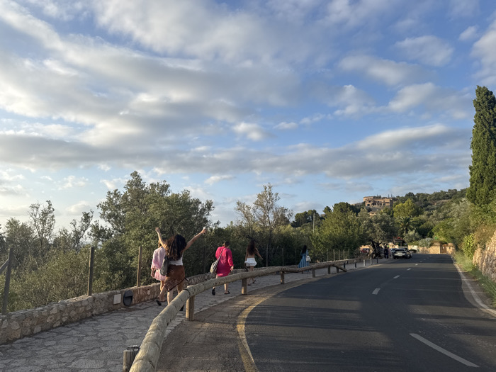 The image displays a curving asphalt road and an adjacent stone-paved sidewalk in Deià, Spain, observed under a blue sky with scattered white clouds. The road, featuring a solid white edge line and a broken white center line, extends into the distance curving towards the right. Five individuals are present on the left-hand sidewalk, walking away from the viewer. The closest person, from behind, has long dark hair, is wearing a white top and a brown patterned skirt, and has both arms raised and extended outwards. Behind this individual, a person in a light pink top and another in a bright pink top are visible. Further back on the sidewalk, a fourth person wears a white top and patterned skirt, and a fifth wears a blue patterned dress. The sidewalk is bordered by a low dry-stone wall and a wooden post-and-rail fence. The surrounding environment consists of lush green Mediterranean trees and bushes covering rolling hills. Several terracotta-roofed buildings are nestled among the foliage on the distant hills. A tall, dark, columnar tree stands prominently on the far right side of the road. In the distance on the right side of the road, a white SUV is parked or stopped, with two indistinct figures standing near it. The lighting suggests late afternoon or early evening.