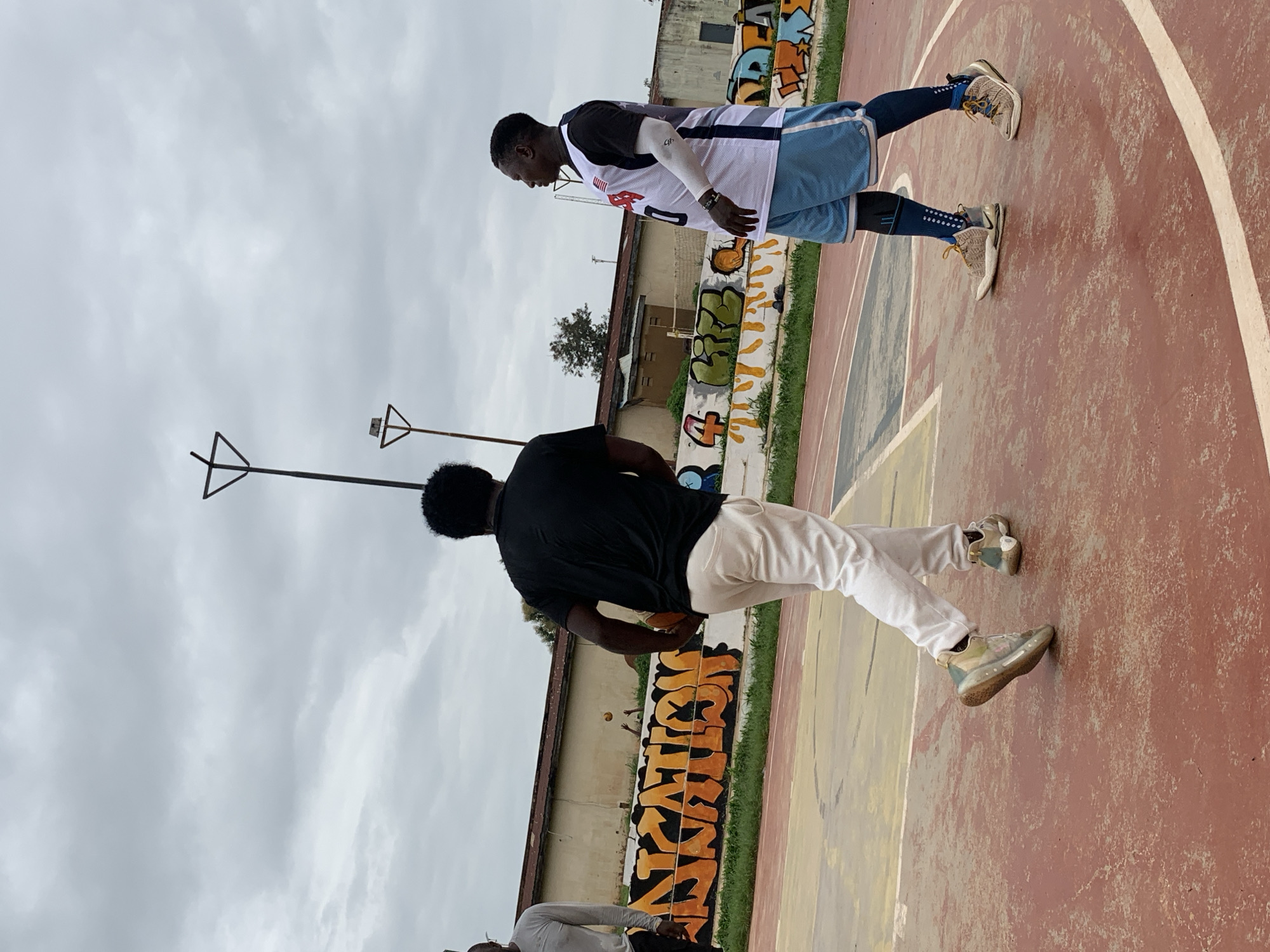 Two men are playing basketball on an outdoor court in Jos, Nigeria. The court surface is red with painted lines. A wall covered in orange and green graffiti is visible in the background, along with a building and trees. The sky is overcast. One man, wearing a black t-shirt and white pants, is holding a basketball and facing away from the camera. The other man, wearing a white and blue basketball jersey, blue shorts, and a white arm sleeve, is facing the first man. A third person is partially visible on the left, wearing a grey long-sleeved shirt. Two metal poles with triangular top structures are present on the court.