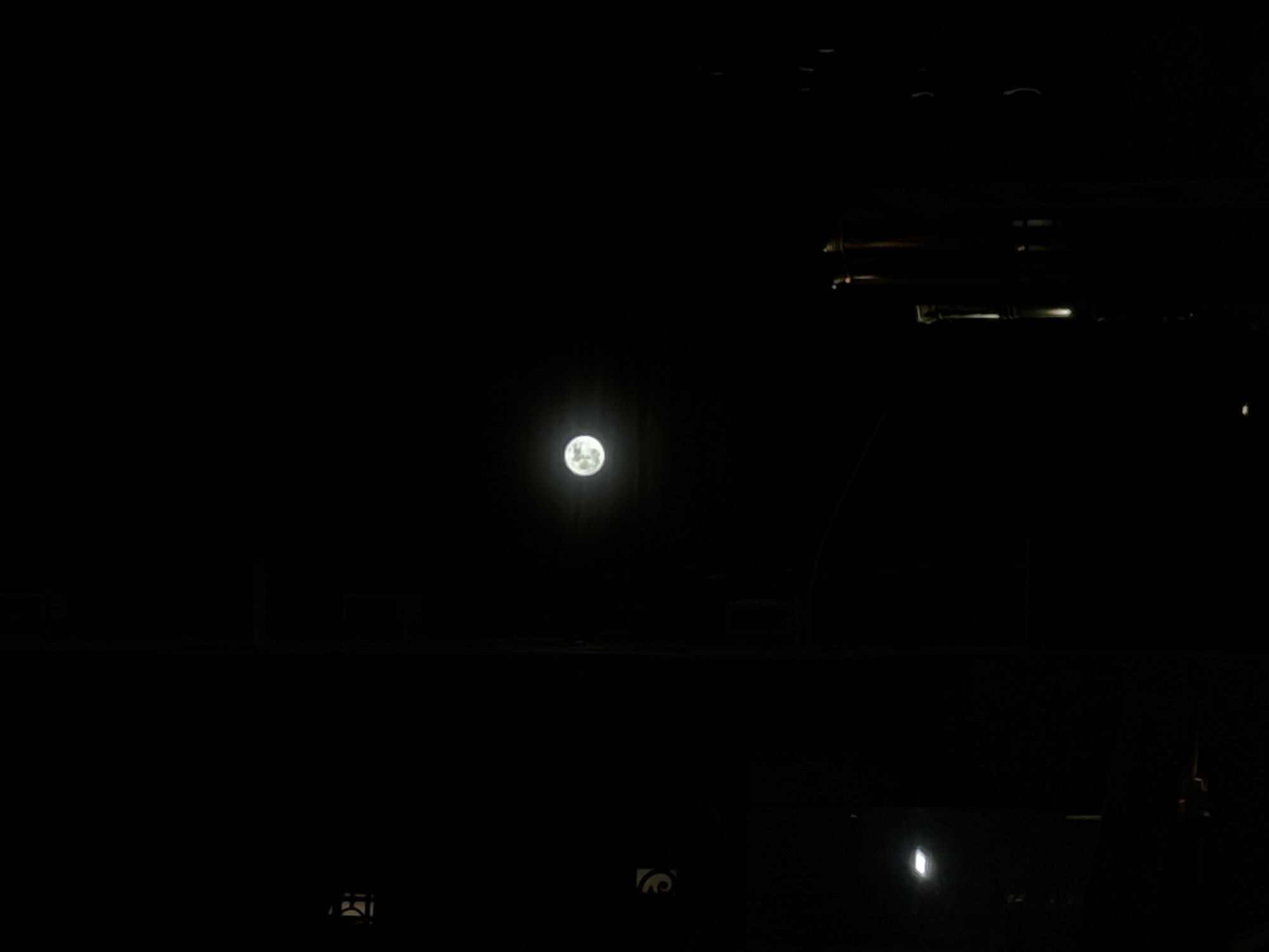 The image depicts a night scene, dominated by darkness, with the moon as the primary light source. The moon is positioned in the center of the frame. In the lower part of the image, details suggest an urban setting, possibly Yerevan, Armenia. Structures and elements like power lines are visible as silhouettes. There are additional light sources that are dim.