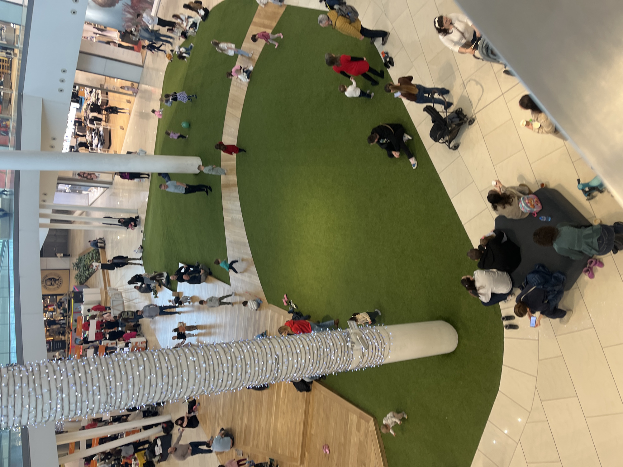 The image displays a multi-level shopping mall interior in Unknown, Unknown. A large, irregularly shaped artificial green turf play area occupies a central common space on the ground floor, bordered by light-colored wooden platforms and a light-tiled walkway. A prominent white cylindrical column, decorated with string lights, stands within the turf, extending upwards through visible upper levels.
Approximately fifty individuals are present across the visible areas. Adults are predominantly seated on the wooden platforms or at the edge of the turf, supervising, conversing, or using mobile phones. Several adults are also standing, observing the play area. Children, ranging from toddlers to elementary school age, are actively playing on the artificial turf, observed crawling, walking, running, and interacting with a green ball. Some children are seated with adults.
Objects visible include strollers parked on the tiled walkway, footwear placed at the edge of the turf, and personal bags. Retail storefronts are visible on multiple levels in the background, including one with a 'Five Guys' restaurant sign. The environment is brightly illuminated by overhead artificial lighting.