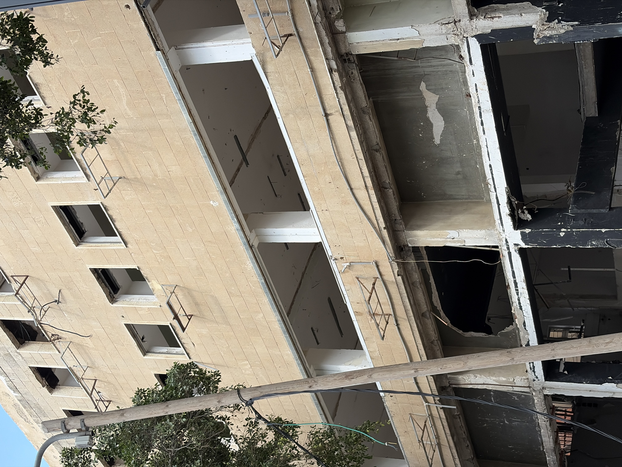 The image shows a damaged building in Tel-Aviv, Israel. The building is multi-story and appears to be in a state of disrepair, with missing windows and exposed interior spaces. The building is constructed of a light-colored stone or concrete. The interior spaces visible through the openings in the building show bare walls and ceilings, with evidence of damage and deterioration. Electrical wires and conduit are visible running along the exterior of the building. A utility pole and tree branches are in the foreground.