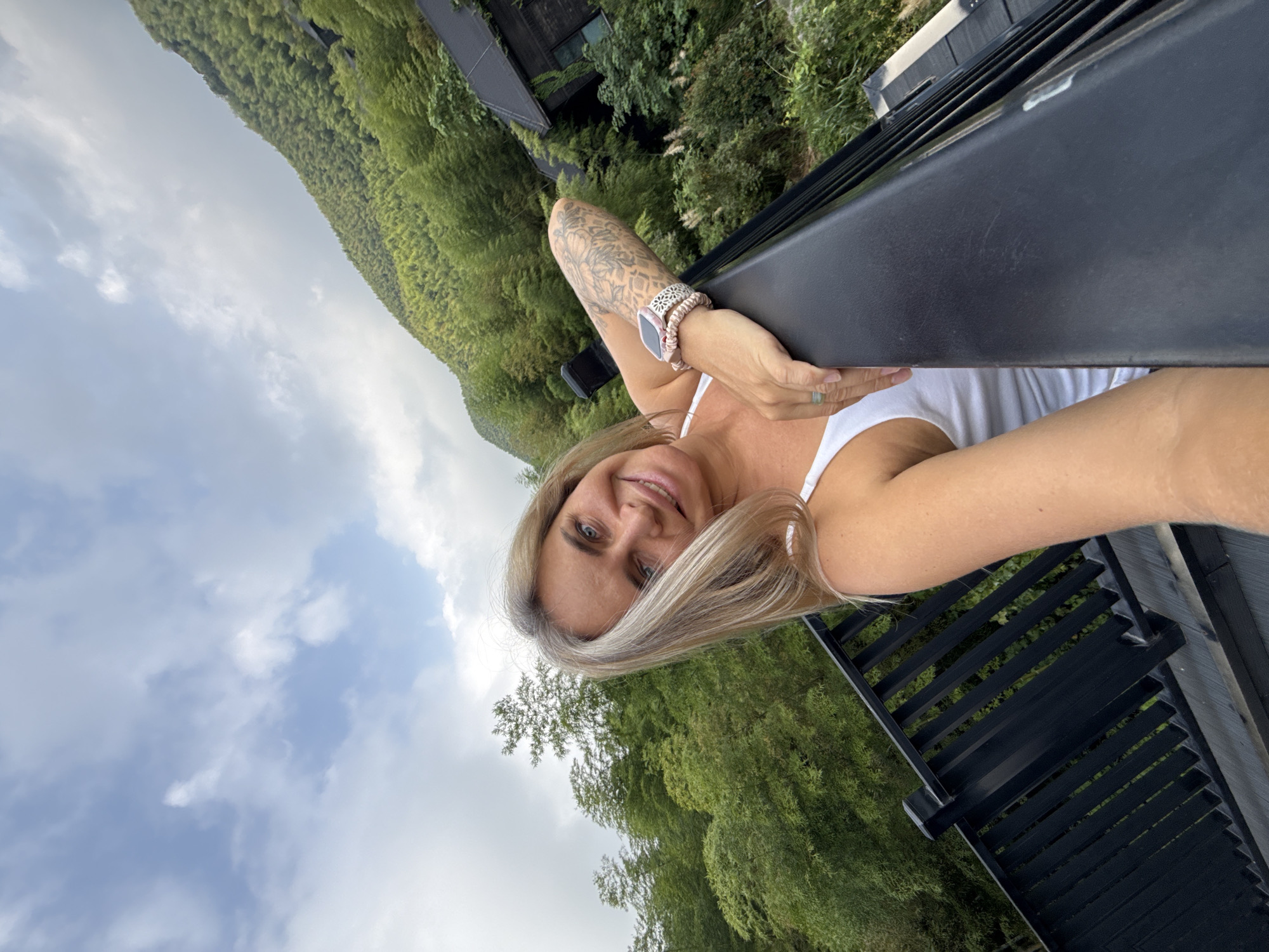 A woman with blonde hair smiles at the camera, leaning on a black railing. She is wearing a white tank top and a watch on her left wrist. Behind her, lush green trees and a hillside are visible under a cloudy sky. A dark, modern-looking building is nestled into the hillside. The setting appears to be a mountainous area with dense vegetation, possibly in the city of Wuxing District, China, given the style of architecture and the natural landscape.