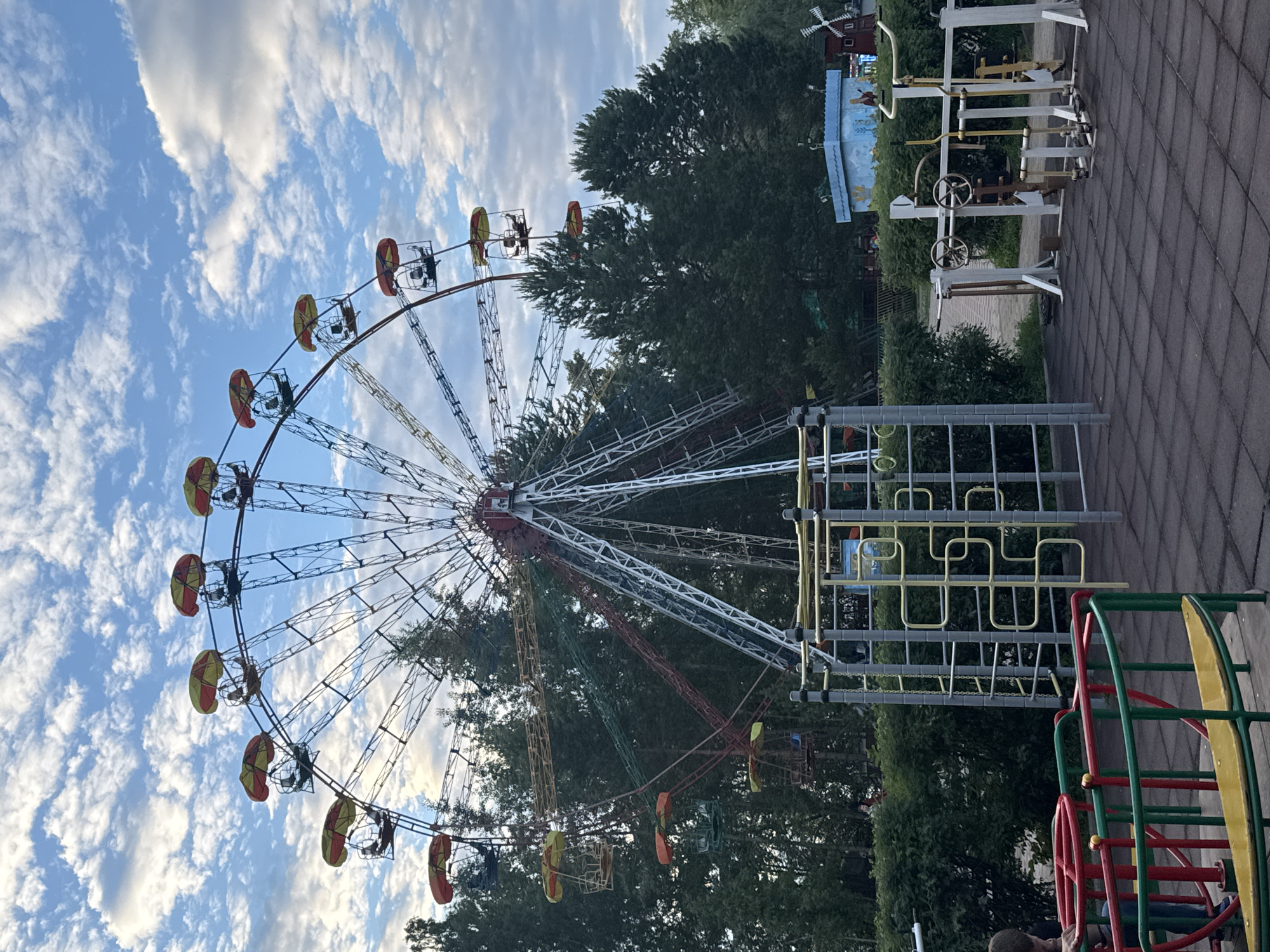This image depicts a Ferris wheel and a playground in Chaikovsky, Russia. The Ferris wheel is large and appears to be made of metal with several colorful gondolas, each topped with a red and yellow umbrella-like canopy. The wheel is set against a backdrop of trees and a cloudy sky. In the foreground, there is a playground area with various pieces of equipment, including a merry-go-round with red and green railings and yellow seating, a jungle gym with yellow bars, and several outdoor fitness machines. The ground in the playground is covered with dark, square rubber tiles. In the background, there are more trees and a small blue building with a painted mural, along with a miniature windmill. There are no people clearly visible in the image, though a person's arm and shoulder can be seen on the left side, interacting with the merry-go-round. The overall scene suggests a public park or amusement area.