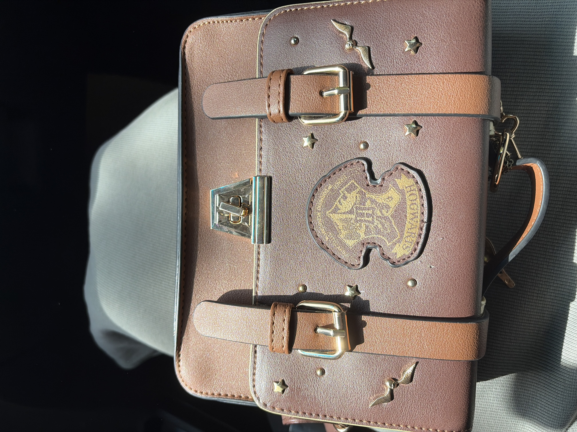 A close-up shot depicts a brown, faux-leather bag with a Harry Potter theme. The bag features two decorative straps with gold-colored buckles, a gold-colored latch, and a patch of the Hogwarts crest. There are also gold-colored star and winged-key decorations. The bag is resting on what appears to be a light gray fabric surface. The location and existence of the city of Chaikovsky, Russia are not evident in the image.