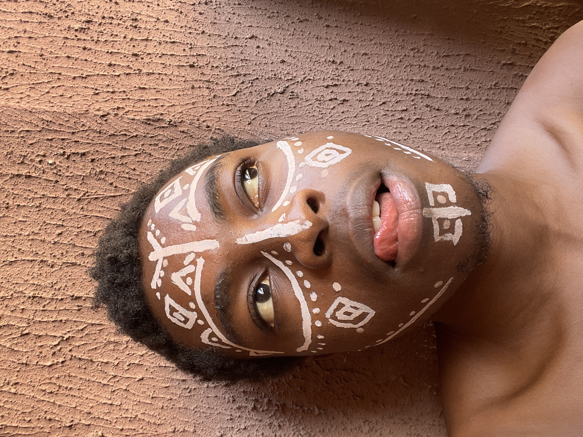 A close-up, low-angle shot shows a Black person, likely male, with white tribal-style face paint lying against a rough, textured brown surface. The person's head is tilted, and they are looking upward with their mouth slightly open, tongue visible. The texture of the surface beneath the person is reminiscent of a coarse, earthen material. The location in the city of Nkpor, Nigeria, is not verifiable from this image.