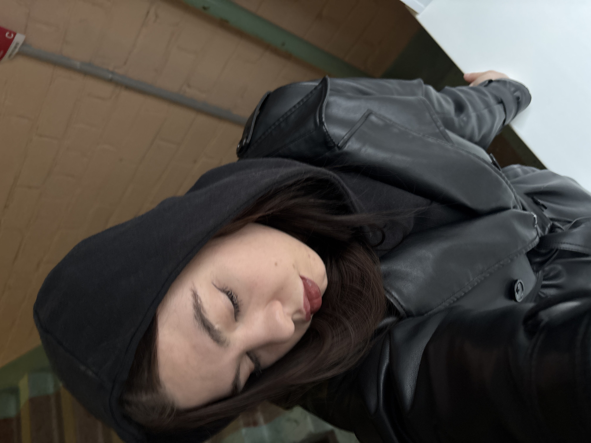 The image, rotated 90 degrees clockwise, presents a close-up of a person, likely female, from a slightly low-angle perspective. The person's head is positioned towards the left of the frame. They have fair skin, dark eyebrows, and dark brown hair partially covered by a black hooded sweatshirt. Their eyes are closed, and their lips are slightly parted, revealing a subtle hint of red lipstick. The person is wearing a black leather jacket over the hoodie, with visible details like seams and a button on the jacket.
The background on the left side of the frame features a light brown brick wall. A grey pipe runs horizontally across this wall, and above it, a small red object with a white letter "C" is partially visible, suggesting a sign or part of an electrical or safety system. Further right and slightly above, a hint of a green beam or pipe can be seen. On the right side of the image, a large, plain white surface is visible, possibly a desk or wall, against which the person's arm, clad in the black leather jacket, appears to be resting.
The scene appears to be indoors under ambient lighting, offering no specific clues about the time of day or weather. The closed eyes and relaxed expression suggest a moment of rest, contemplation, or a posed selfie.