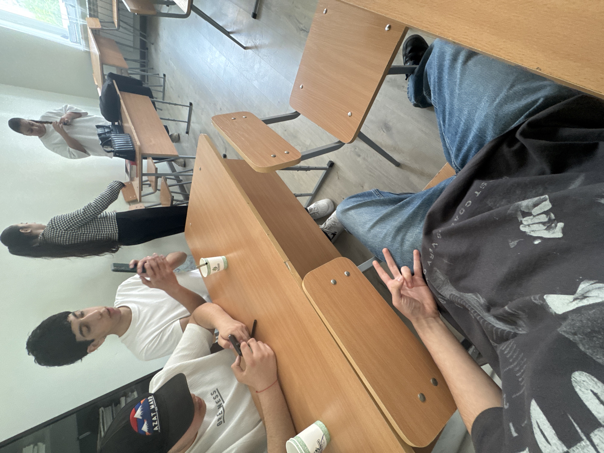 This image, taken from a high, angled, and rotated perspective, depicts an informal scene inside what appears to be a classroom or lecture hall in Yerevan, Armenia. Several young individuals are seated at light-wood desks with grey metal frames.
In the foreground on the right, the legs of a person wearing blue jeans and a dark t-shirt are visible. Their left hand is raised, displaying a three-finger 'peace' or 'W' gesture.
Towards the center-left, two people are seated at a desk. The one closest to the viewer wears a white t-shirt with "GRE NESS" visible on the chest and a black baseball cap emblazoned with "AZAT MANP". They are holding and interacting with a smartphone. Beside them, another person with dark hair, also in a white t-shirt, is similarly engaged with a smartphone. Two paper cups with a green design are placed on the desk in front of them.
Further back on the left side of the frame, a woman with long dark hair, wearing a black and white patterned (houndstooth) top and black pants, is seated at a desk, looking down at her phone. Behind her, another person in a white t-shirt stands near a desk by a large window, appearing to braid or style their long hair. A black backpack rests on the desk next to this individual.
The setting features light-colored walls and a light wooden floor, with natural daylight streaming in from the window, suggesting it is daytime. The overall atmosphere is relaxed, with the individuals largely focused on their personal devices or activities.