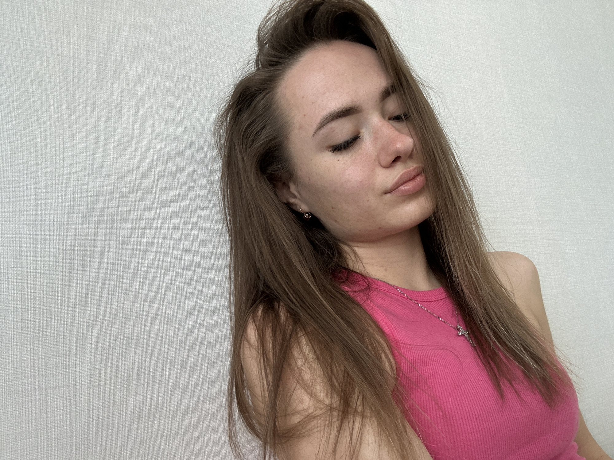 A close-up portrait captures a young woman indoors, positioned against a plain, light gray or off-white textured wall. Her head is tilted slightly, and her eyes are closed, conveying a serene and relaxed expression. Her lips are softly parted in a slight pout. She has long, straight brown hair, styled so that some strands fall across her forehead and frame her face, while a large section rests over her left shoulder and chest.
She is wearing a bright pink ribbed tank top. A silver chain necklace with a small, intricate charm, possibly a cross or a decorative pendant, is visible against her top. In her visible left ear, she wears a small hoop earring adorned with a dark or reddish stone. Her skin shows some natural freckles across her forehead, nose, and cheeks. The lighting is soft and even, suggesting an indoor setting without direct harsh light. No specific location cues beyond the wall texture are visible, and there is no discernible text in the image.