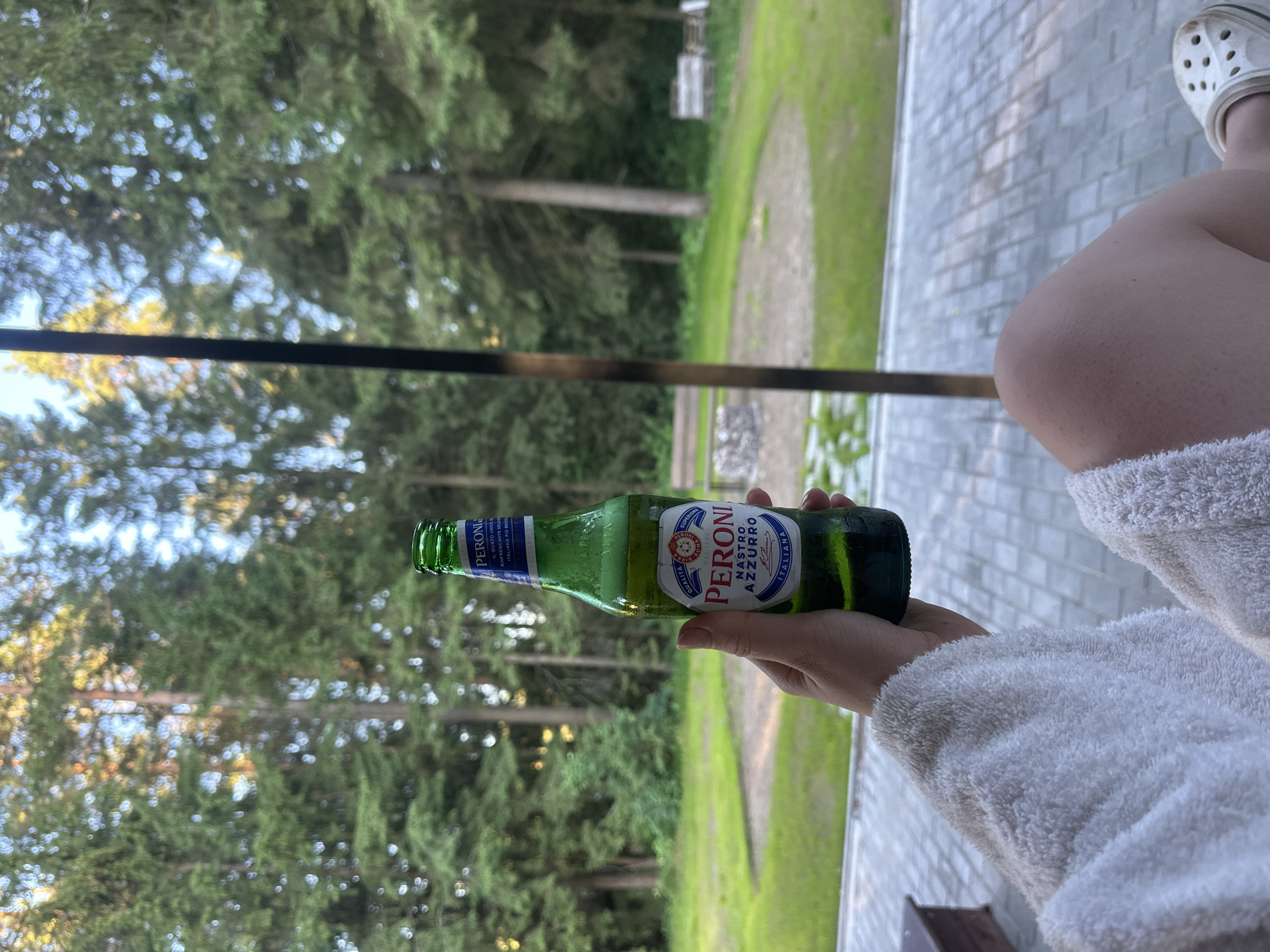 A person's hand, wearing a white terrycloth robe, holds a green glass bottle of Peroni Nastro Azzurro beer. The bottle is partially obscured by the robe and the person's leg, which is also visible, wearing a white Croc shoe. The background shows a natural outdoor setting with tall evergreen trees, a grassy area with a fire pit, and a paved patio. The lighting suggests it is daytime, possibly late afternoon or early evening. The location is described as Priazhka, Russia.