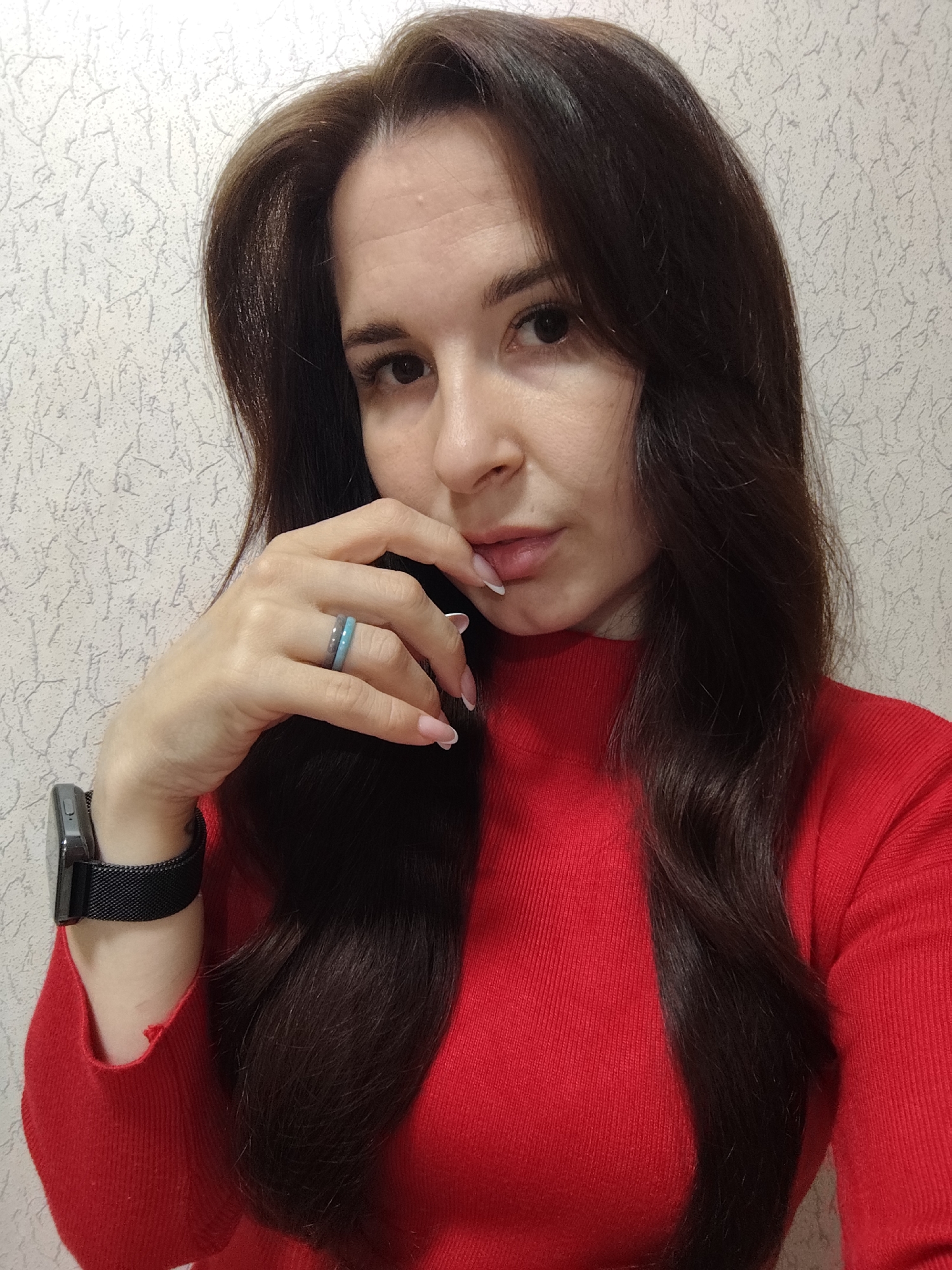 A young woman is captured in a close-up, chest-up selfie, looking directly at the camera with a contemplative expression. She has long, dark brown, wavy hair that falls over her shoulders. She is wearing a fitted, ribbed red turtleneck sweater. Her right hand is raised, with her fingers lightly touching her lips, showcasing a French manicure on her fingernails. On her right ring finger, she wears two thin rings, one appearing grey and the other a light blue or teal color. A smartwatch with a black mesh band is visible on her left wrist. The background is a light-colored, possibly white or light grey, textured wallpaper with a subtle, abstract, vine-like pattern. The lighting appears even, suggesting an indoor setting. The image was taken in Fokino, Russia. There is no visible text.