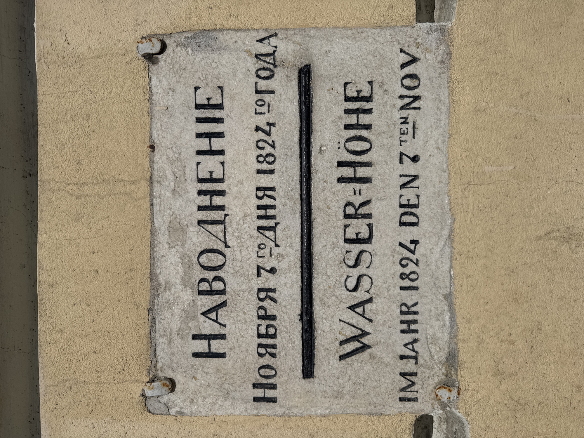 A close-up photograph captures a historical rectangular stone plaque embedded in a light beige stucco wall, likely in Saint Petersburg, Russia. The plaque, made of light gray, textured stone, commemorates a significant flood. It features inscriptions in two languages, Russian (Cyrillic script) on the left and German (Latin script) on the right, separated by a distinct vertical black line representing the high water mark.
The Russian text on the left reads:
"НАВОДНЕНІE" (Navodnenie), which translates to "Flood."
Below that, "НОЯБРЯ 7 ДНЯ 1824 ГО ГОДА" (Noyabrya 7 dnya 1824 go goda), meaning "November 7th day 1824 of the year."
The German text on the right reads:
"WASSER-HÖHE," which translates to "Water-Height" or "Water-Level."
Below that, "IM JAHR 1824 DEN 7 TEN NOV," meaning "In the year 1824 the 7th Nov."
The plaque is affixed to the wall with visible, old metal fasteners in its corners, some of which show signs of rust and wear. The surrounding wall exhibits minor cracks and a weathered texture. The lighting indicates an outdoor setting, but specific time of day or weather conditions are not discernible. The plaque serves as a historical marker, documenting the water level during the notable flood of November 7, 1824.