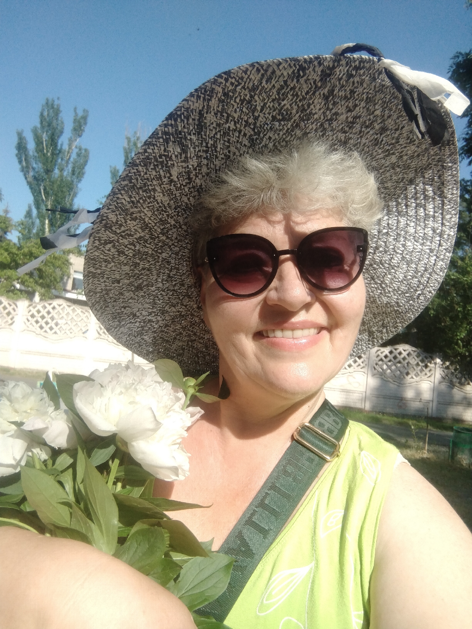 A woman with short, gray hair, wearing large, dark sunglasses and a wide-brimmed, patterned hat, smiles while holding a bouquet of white peonies. She is wearing a light green, sleeveless top with a white leaf pattern and a green strap across her chest with gold hardware. The background suggests an outdoor urban setting with trees, a white decorative fence, and a clear blue sky. The scene is likely in the city of Horishni Plavni, Ukraine, as indicated by the user prompt.