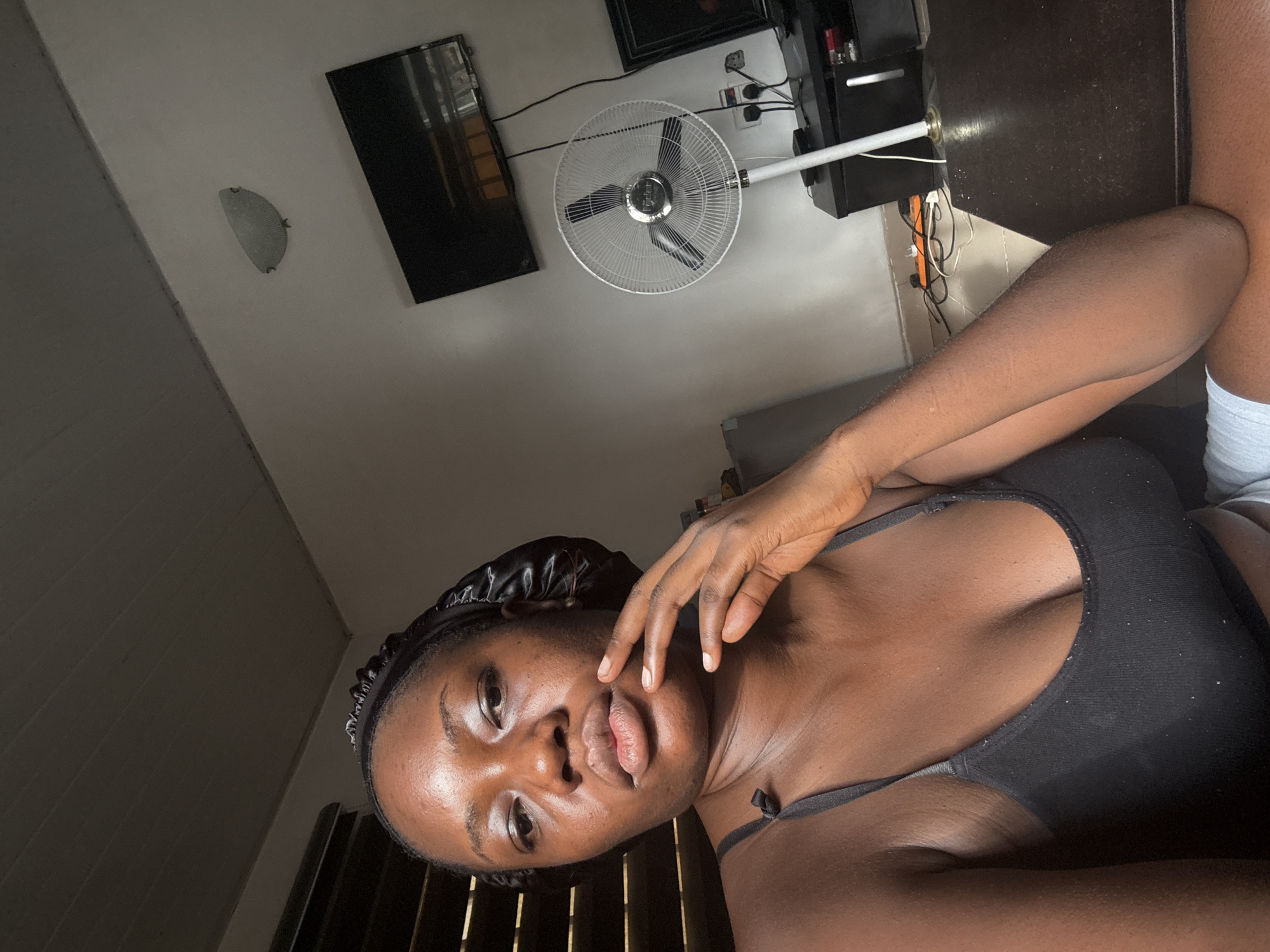 A woman lies indoors. She wears a black satin bonnet and a black bra. A standing fan, a television, and a wall-mounted light fixture are visible in the background. The location appears to be indoors, possibly a residential space. The location is not specifically identifiable as being in Abuja, Nigeria from the image alone. The image depicts a single individual posing for a selfie, with no obvious actions or interactions with others visible.