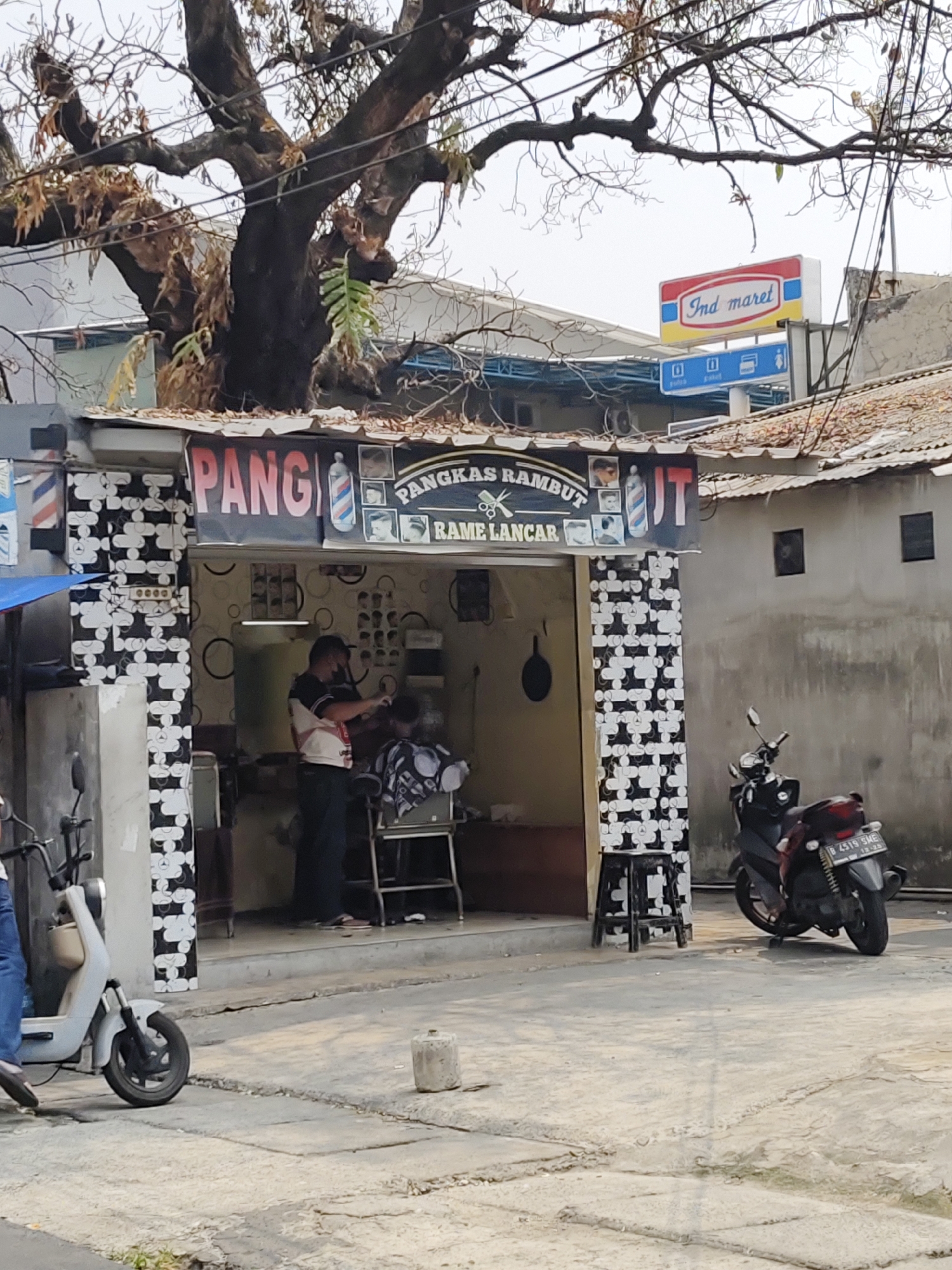 The image depicts a street scene in South Jakarta, Indonesia, featuring a barbershop and its surroundings. In the foreground, a light-colored electric scooter is parked to the left, with a person wearing blue jeans and dark shoes visible on the left side of the frame, their lower legs and feet near the scooter. To the right of the scooter, the street surface is cracked concrete.
The central focus is a barbershop with a sign that reads "PANGKAS RAMBUT RAME LANCAR" in red and black lettering, adorned with images of haircuts and barber poles. The barbershop itself is open, revealing a barber wearing a mask, a white and red striped shirt, and dark pants, actively cutting the hair of a customer seated in a chair. The interior of the shop is decorated with a patterned wallpaper and features several barber chairs and tools.
To the right of the barbershop, a dark-colored motorcycle is parked on the street. The background shows a building with a tiled roof and gray walls, and above it, a prominent Indomaret convenience store sign. A large, old tree with bare branches and some dry leaves extends over the barbershop and the street. The overall environment suggests an urban street in a commercial area.