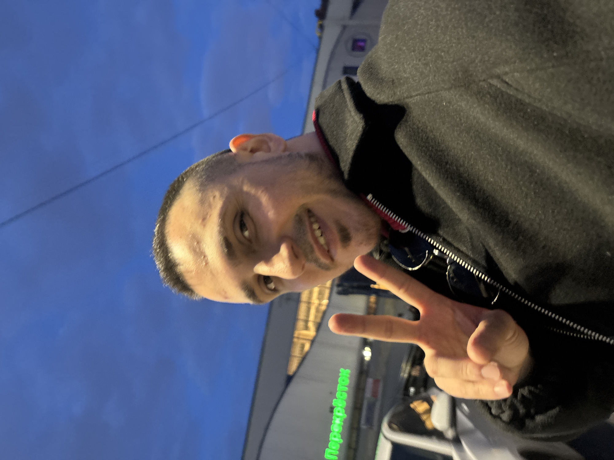A man is posing for a selfie outdoors at dusk in Saint Petersburg, Russia. He is giving a peace sign with his right hand and smiling at the camera. He has a shaved head and a short beard. He is wearing a dark-colored coat with a zipper visible and a red collar.
In the background is a building with the word "Перекресток" (Perekrestok) written in green, Cyrillic letters. Perekrestok is a supermarket chain in Russia. The building appears to be lit up, with several lights visible in the background. The sky is a blue color, indicating either dusk or dawn.
There are cars visible behind the man, and the overall setting suggests he is in a public area, possibly near a supermarket.