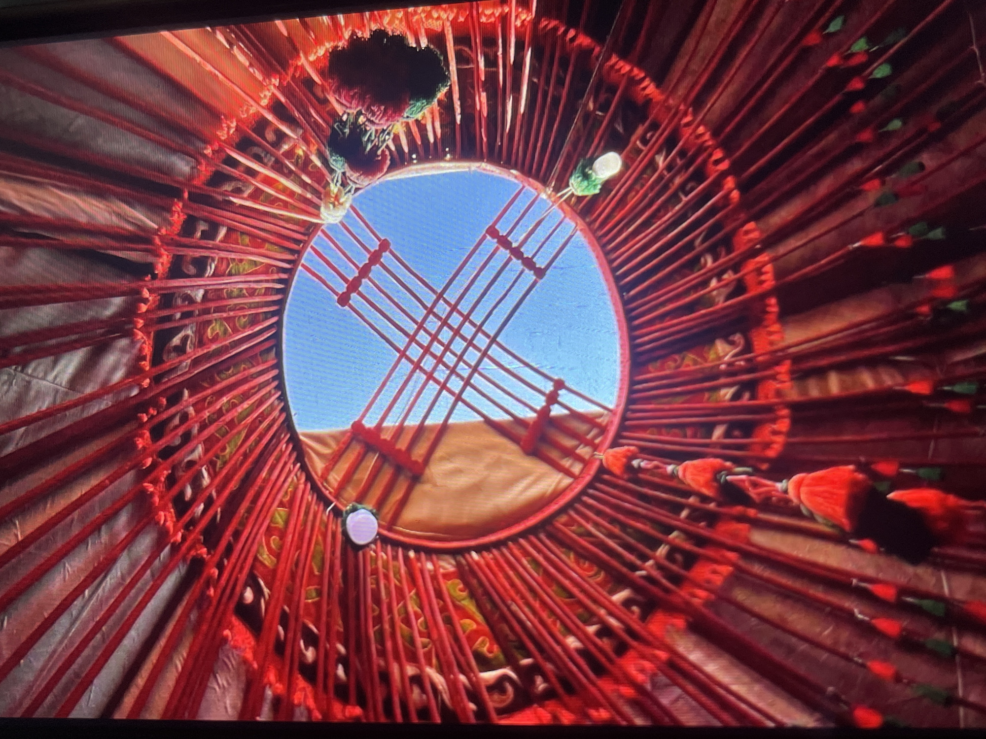 The image shows the interior of a yurt, a traditional dwelling of nomadic peoples of Central Asia, likely in Bishkek City, Kyrgyzstan. The viewpoint is from inside the yurt looking upwards towards the circular opening at the top, known as the *tunduk*. The structure is characterized by a lattice of wooden poles radiating outwards and upwards, all wrapped in vibrant red fabric or wool. Decorative tassels, in shades of red and green, hang from various points along the structure, adding a festive touch. The *tunduk* reveals a clear blue sky, suggesting it is daytime and the weather is fair. The woven interior walls of the yurt are visible, adorned with colorful patterns, hinting at the cultural significance and artistry associated with these dwellings. There are no people or specific actions depicted; the focus is solely on the architectural and decorative elements of the yurt's interior.