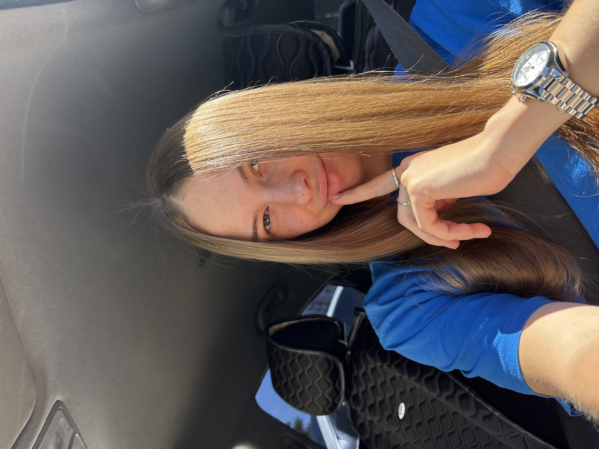 A young woman with long, blonde hair is pictured inside a car, taking a selfie. She is wearing a bright blue t-shirt and has her right index finger resting on her lips. She is also wearing a silver ring on her left hand and a silver watch on her left wrist. The interior of the car is visible, including a black headrest with a patterned cover and a seatbelt. Sunlight streams in from the right side of the frame, illuminating her hair and face. The background shows the interior of the car and a window with a view of what appears to be an outdoor environment. There is no specific visual information that can tie this image to the city of Perm, Russia.