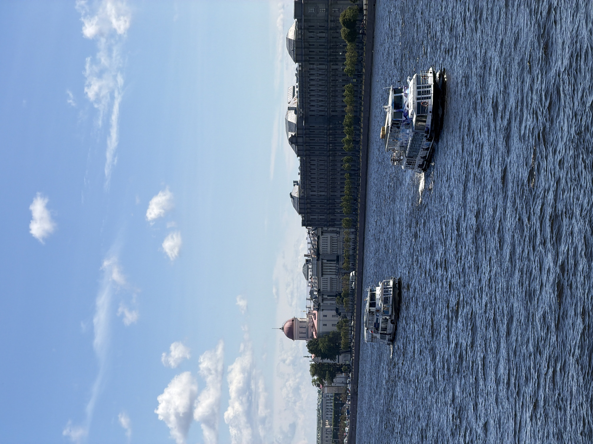 This is a vertical, full shot of the Neva River in Saint Petersburg, Russia. The sky is a bright blue with scattered white clouds, suggesting a sunny day. The water of the river is a deep blue and appears choppy, with small waves rippling across its surface.
Two white passenger boats are visible on the river. The boat in the foreground is closer to the viewer and appears to be a tour boat with multiple decks. The second boat is further in the distance, also on the river.
Along the riverbank, a line of grand, historic buildings forms the cityscape. These buildings are predominantly grey and feature classical architecture with arched windows and ornate facades. Trees line the riverbank in front of the buildings. A prominent pink building with a domed roof is visible amidst the grey structures. The scene depicts a leisurely day on the river, likely a popular time for sightseeing in Saint Petersburg, given the presence of the tour boats. The lighting suggests it is daytime, likely midday given the brightness of the sun. No text is visible in the image.
