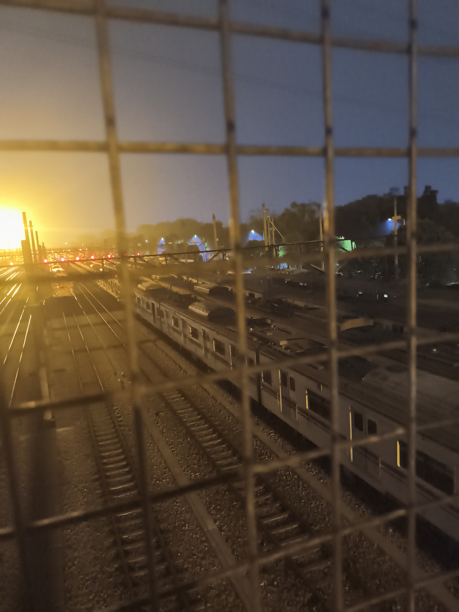 The image is a view of a train yard at dusk or night, seen through a wire fence. The train yard has multiple sets of tracks and several trains are visible, some appearing to be stationary and others possibly in motion. The lighting is a mix of warm, artificial lights from the yard and a muted, natural twilight sky. In the background, there are some trees and structures, suggesting an urban or semi-urban environment. The fence in the foreground is out of focus, creating a blurred effect that frames the scene. The location is identified as Depok, Indonesia.