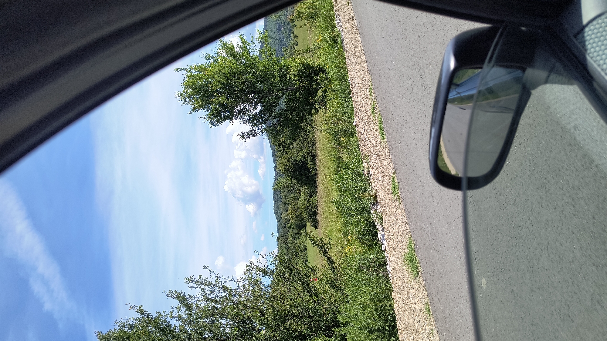 This is a photograph taken from inside a vehicle, looking out of the passenger window. The perspective shows a roadside scene on a clear day. In the foreground, the asphalt road curves to the right, with a gravel shoulder to its left. A side mirror of the car is visible in the lower right portion of the frame, reflecting a portion of the road and the car's interior.
Beyond the road and shoulder, a grassy field stretches into the distance, dotted with wildflowers. Several trees, with lush green foliage, are scattered throughout the landscape. In the background, rolling hills covered in dense forest are visible under a bright blue sky with scattered white clouds. The overall impression is one of a rural landscape during daylight hours. The location is identified as Dramsha, Bulgaria.