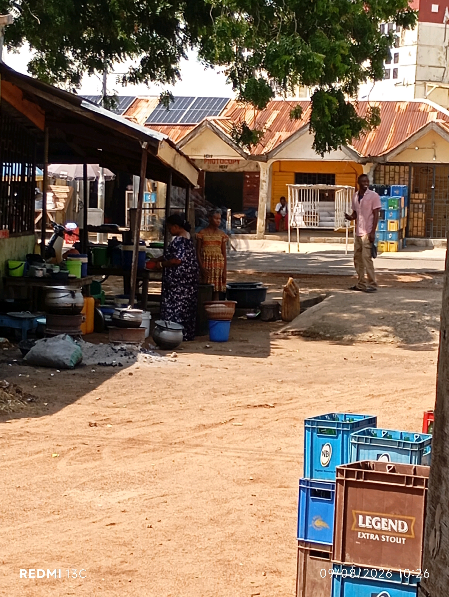 This is a daytime, eye-level, medium shot of an outdoor market scene in Shendam, Nigeria. The foreground is a sandy, uneven surface. To the right, a stack of blue and brown plastic crates, one labeled "LEGEND EXTRA STOUT," is visible. In the mid-ground, under a rustic, sloped wooden awning, two women are interacting near a collection of cooking pots and buckets. One woman, in a dark floral dress, appears to be tending to the cooking area. The other woman, in a patterned dress, stands further back. Behind them, a man in a pink shirt and khaki pants stands near a cage-like structure. Further in the background, buildings with corrugated metal roofs, some equipped with solar panels, line a street. A tree with green foliage partially obscures the upper portion of the scene. The overall impression is that of a bustling, functional market in an African city.