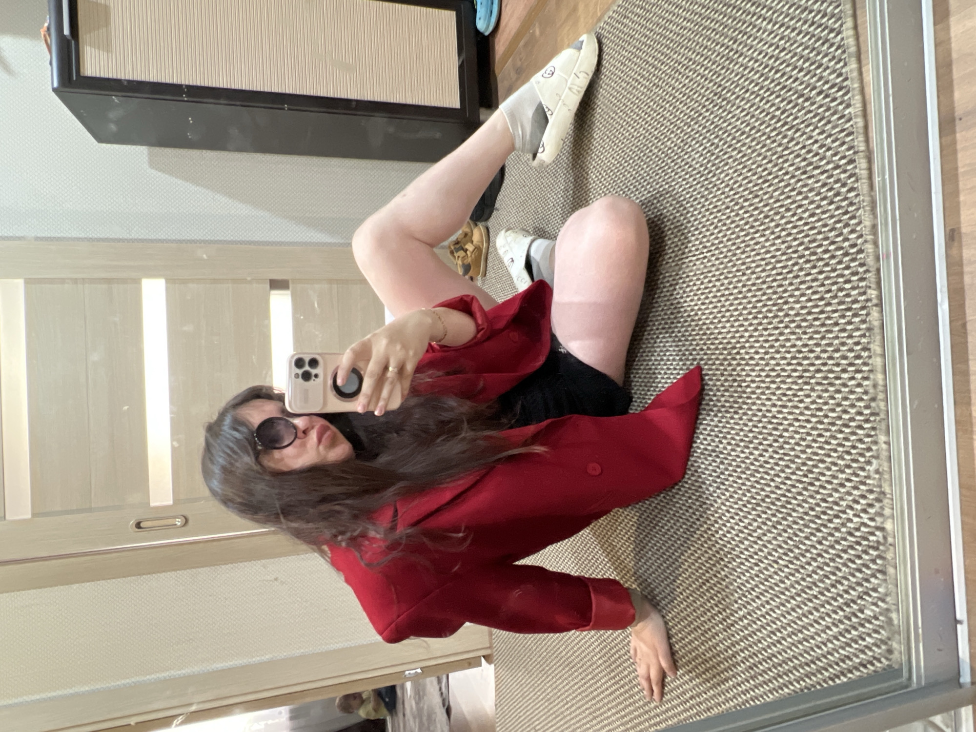 A woman is sitting on a brown and beige textured rug. She is wearing sunglasses and a red coat, taking a photo with her smartphone. Her right leg is extended with her foot in a white sock and patterned slipper. Another shoe can be seen near her left leg, along with a piece of furniture in the background. The scene appears to be indoors, possibly in a room with a mirror. Noviy, Russia is not identifiable within the image context.