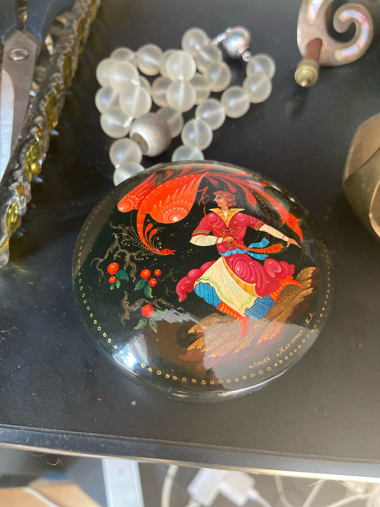 The image shows a dark, reflective surface with several objects. In the center is a round, black lacquered box with a vibrant painting of a woman in traditional Russian attire, possibly depicting a folk tale. The painting features red and orange hues, a woman with flowing hair and a patterned dress, and a stylized bird or phoenix. The edge of the box is decorated with a border of small gold circles. To the left of the box are some metallic scissors with yellow accents and a string of translucent, bead-like objects. To the right, a portion of a gold-colored object is visible. The background is dark and out of focus, suggesting it may be a shelf or table. The text on the box is in Cyrillic script. The objects are arranged in a still life composition, with no discernible activity. The scene does not provide any clues about its location in Kassel, Germany.