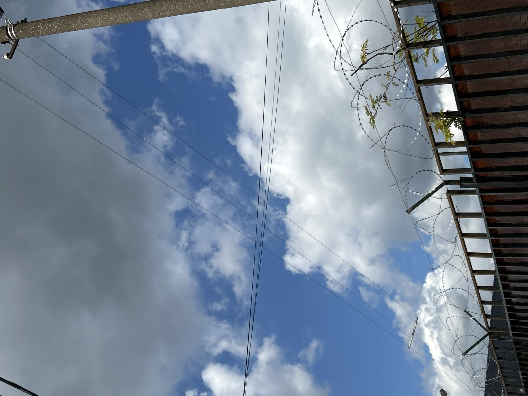 The image shows a sky with clouds, power lines, a concrete utility pole, and a fence with barbed wire. The fence is made of metal posts and wooden planks. There is some greenery growing on the fence. The sky is partly cloudy with blue sky visible. The image is taken from a low angle looking upwards. There are no people or animals visible in the image. The scene appears to be in an outdoor urban or industrial setting.