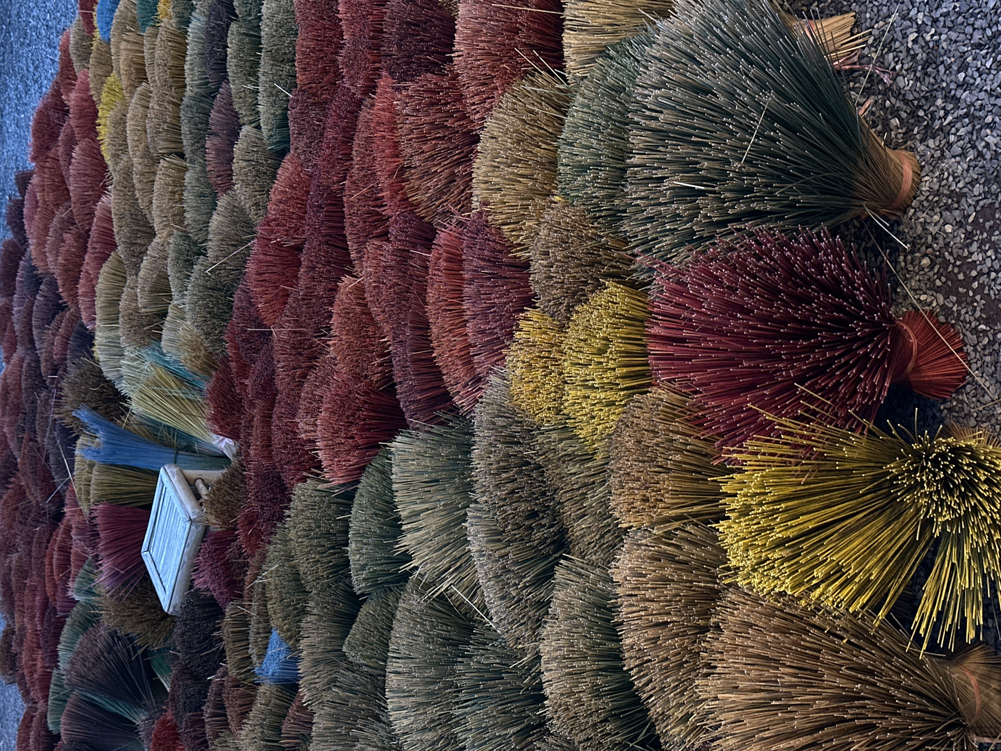The image shows a close-up, overhead view of a large collection of incense sticks arranged in rows and fanned out, creating a textured, colorful display. The incense sticks are of various lengths and colors, including deep reds, burgundy, various shades of brown and tan, muted greens, and some vibrant yellows and blues. They are bundled together at one end, with the other ends splayed out like a brush or a fan. The sticks appear to be dry and are laid out on a surface that resembles gravel or small stones.
In the upper left portion of the image, a small, light-colored rectangular object with a textured surface, possibly a piece of wood or plastic, is visible, partially embedded within the incense sticks. A few bundles of incense sticks appear to have pink or red ties binding them. The lighting suggests it is daytime, but there are no strong shadows indicating a specific time of day or any weather conditions. The setting appears to be outdoors, given the gravelly surface.
The arrangement and density of the incense sticks suggest this is a display or a large stock of materials. Given the context of Nha Trang, Vietnam, this could be at a market, a temple, or a production site for incense. The overall impression is one of abundance and a vibrant, tactile presentation of a common cultural item. There is no visible text in the image.