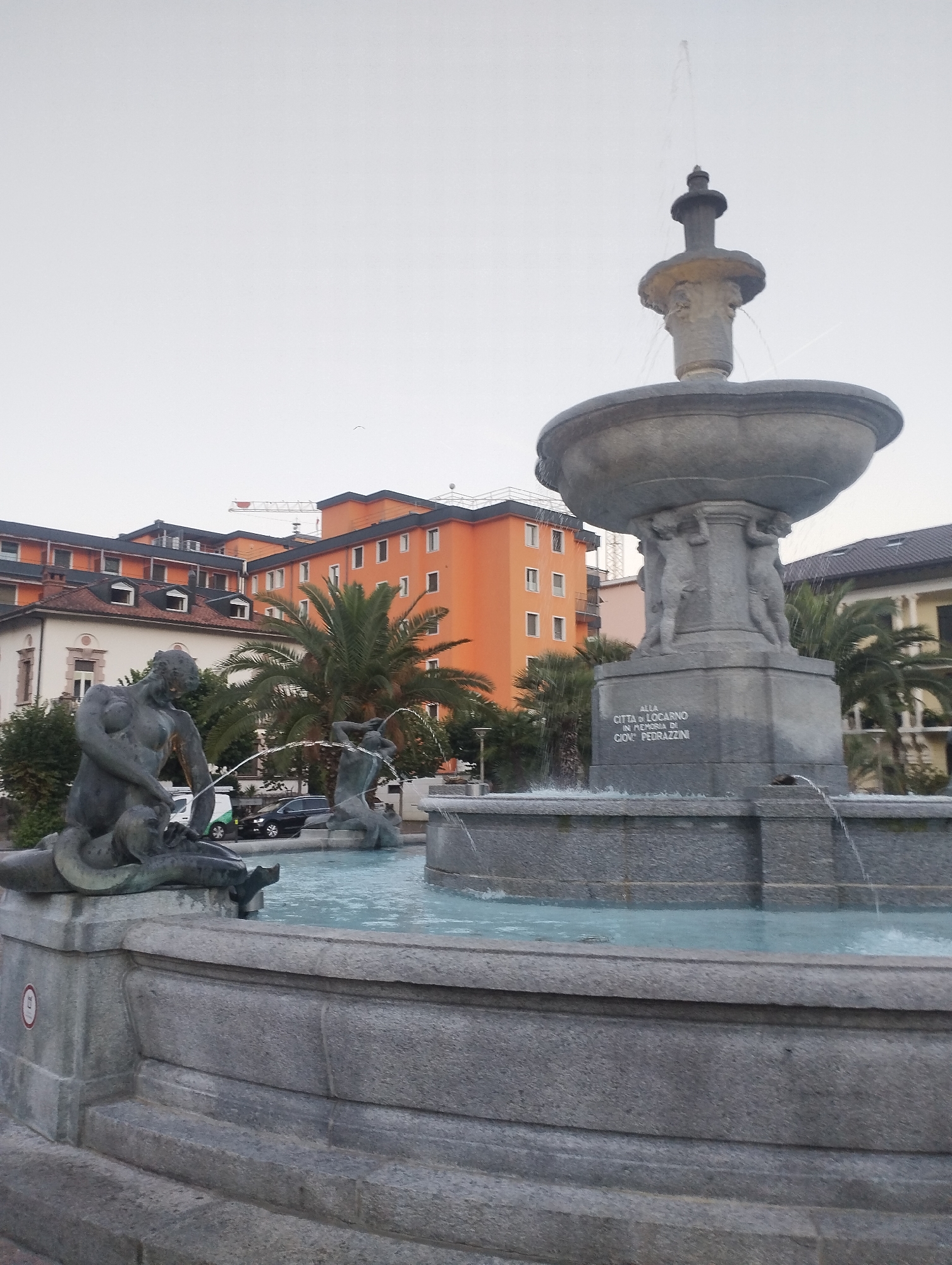 A frontal, slightly low-angle, eye-level outdoor shot captures a stone fountain in a city square in Locarno, Switzerland. The fountain is the central subject, featuring a large basin supported by sculpted figures, with water spouting from its upper levels and cascading into the lower basin. To the left, a detailed sculpture depicts a muscular male figure partially submerged in water, with a smaller figure by his side. Behind the fountain, several buildings are visible, including an orange, multi-story building and a white building with a red-tiled roof. Palm trees and other greenery are interspersed throughout the scene. Cars are parked in the background. The sky is overcast. A plaque on the fountain reads "ALLA CITTA DI LOCARNO IN MEMORIA DI GIOV. FLORAZZINI".
