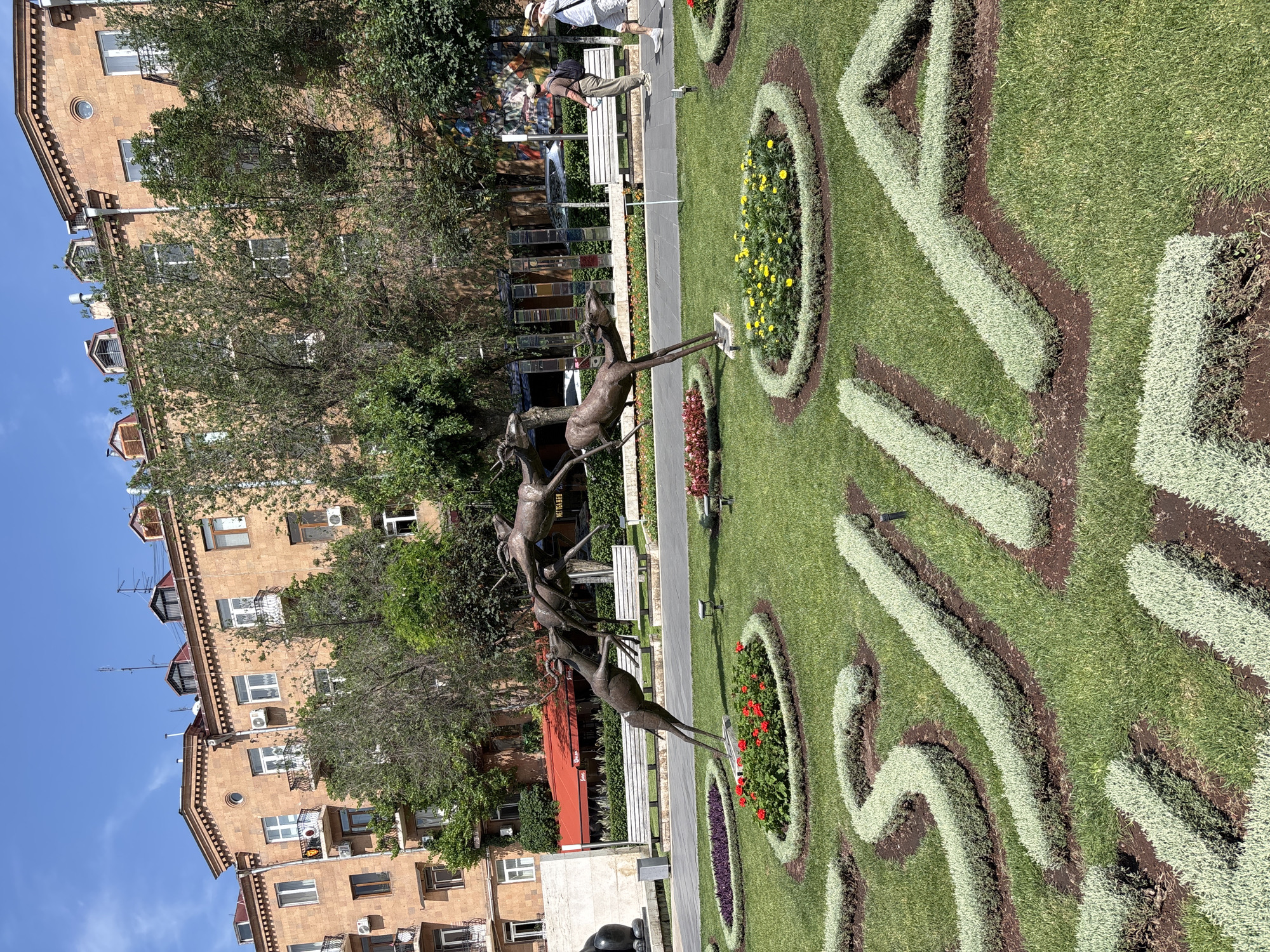 The image captures a sunny day scene in Yerevan, Armenia. In the foreground, a meticulously manicured lawn features a decorative garden design formed by low-lying green bushes, outlining what appears to be the word "YEREVAN". Within the garden beds, circular arrangements of vibrant red and yellow flowers add color to the verdant landscape. Dominating the mid-ground is a dynamic bronze sculpture depicting a group of antelopes in motion, their bodies intertwined as if in a chase or playful interaction. Behind the sculpture, several white park benches are visible, suggesting a public space designed for relaxation. Further in the background, a street lined with trees separates the park area from a multi-story building constructed with light brown stone. The building has numerous windows, balconies with railings, and a distinctive architectural style with decorative elements at the roofline. Two individuals, one wearing a hat and a backpack, and another in a straw hat and shorts, are seen walking on the paved path behind the benches. The sky above is clear blue with scattered white clouds. The overall impression is one of a well-maintained urban park, likely a popular spot for locals and tourists to enjoy the outdoors and appreciate the city's art and architecture.