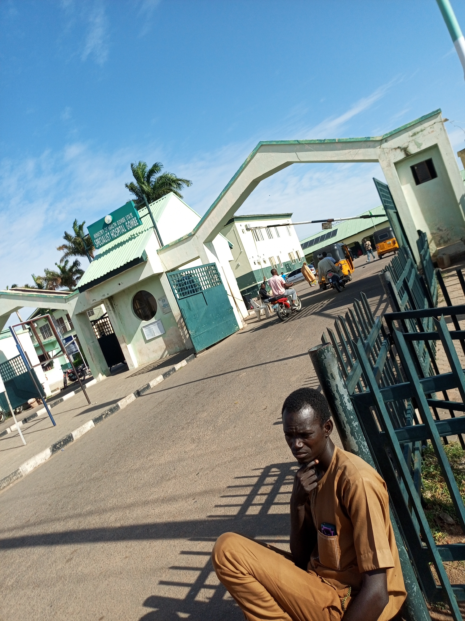 The image shows the entrance to the Specialist Hospital Gombe, located in Arawa, Nigeria. A man in a brown tunic and trousers is sitting on the ground in the foreground, leaning against a green metal fence and touching his chin. Behind him, beyond the entrance gates, is a road with several vehicles, including motorcycles, a yellow tuk-tuk, and a yellow car. People are visible walking and riding the vehicles. The hospital buildings, painted in light green and white, are visible in the background, with a sign reading "MINISTRY OF HEALTH GOMBE STATE SPECIALIST HOSPITAL GOMBE." The sky is blue with scattered clouds. The scene appears to be during the daytime.