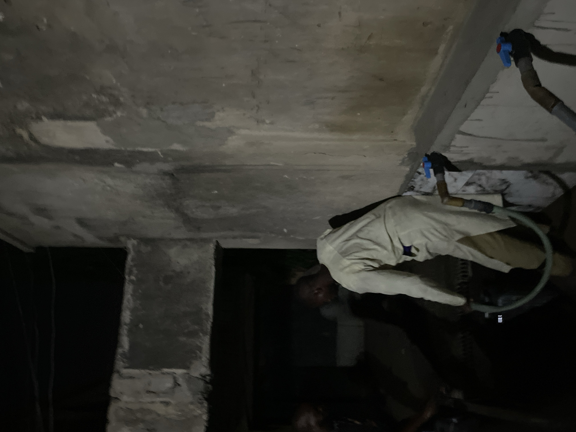 A person wearing a white jacket and tan pants is working with a hose and nozzle system against a concrete ceiling. The room appears to be dark and unfinished, with a concrete pillar and other unseen structures.